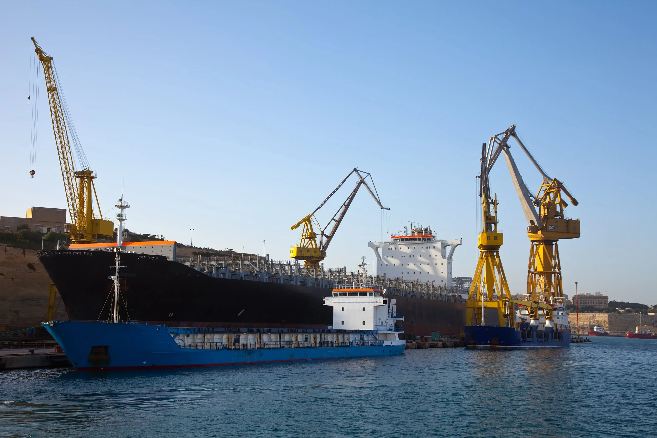 Ships docked at a harbor with yellow cranes and clear blue sky.