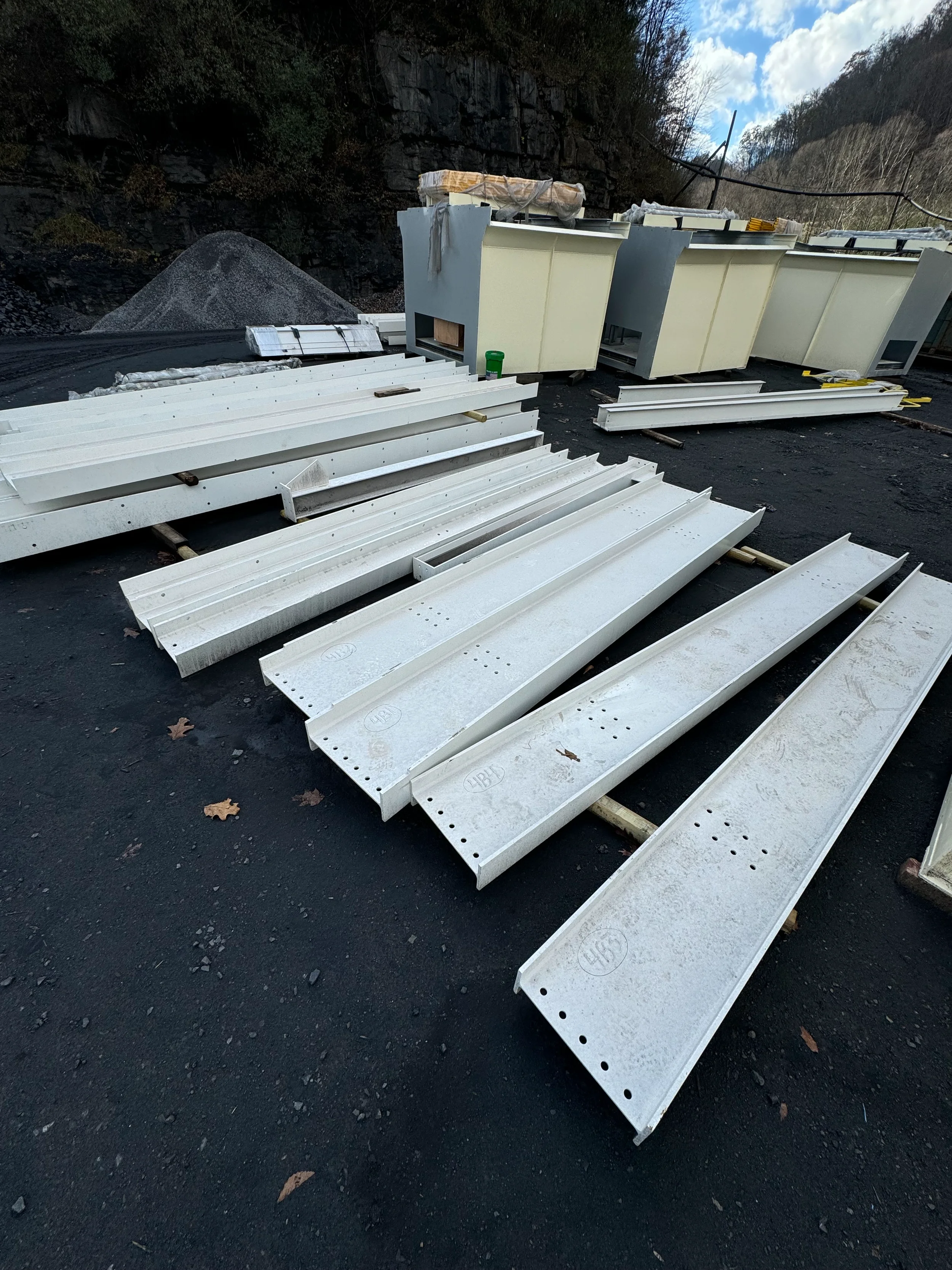Construction materials including metal panels, large cabinets, and other building supplies laid out on a black asphalt surface outdoors, with a rocky hillside in the background and cloudy sky.