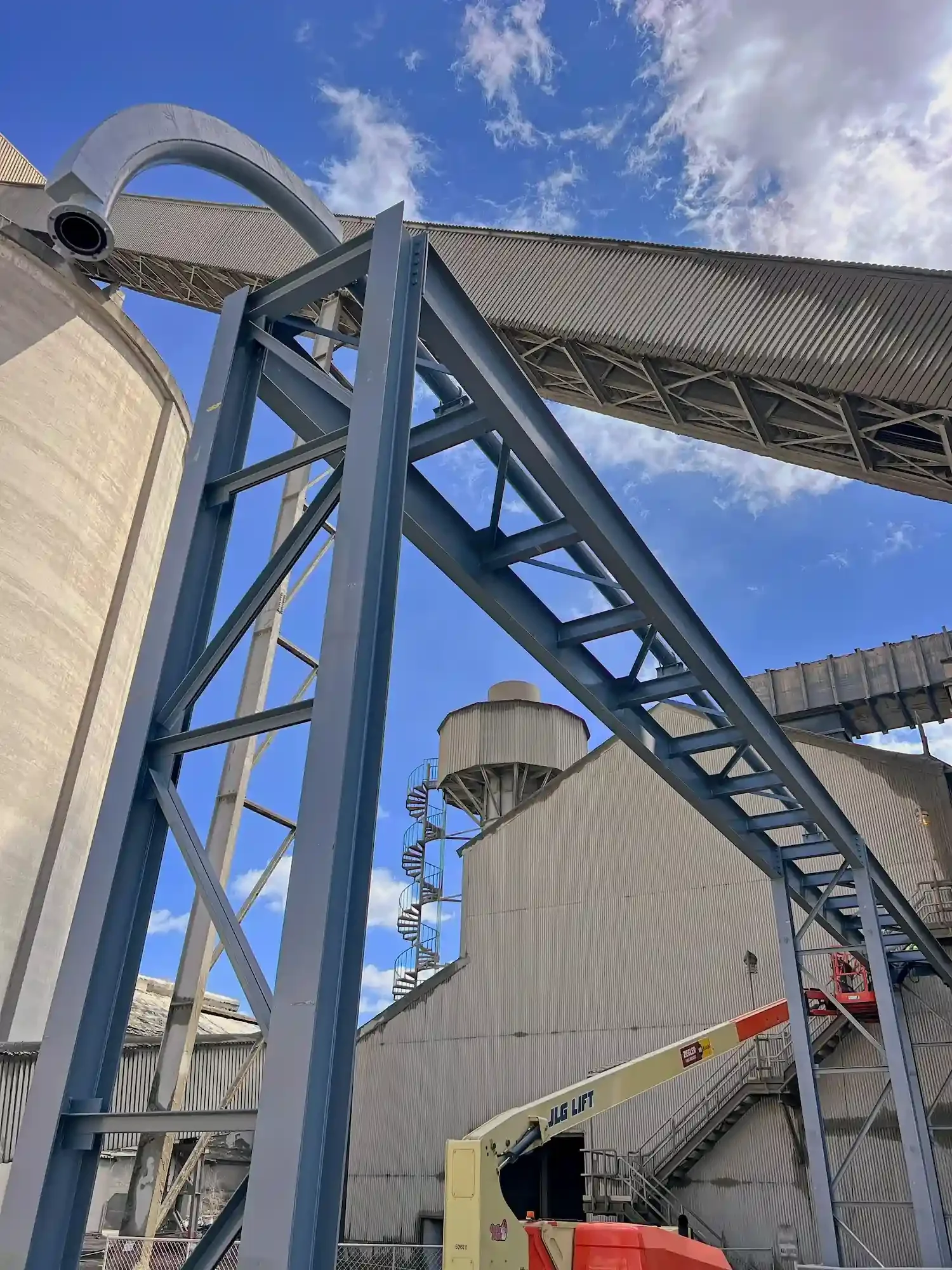 An industrial facility with a blue metal framework and a large cylindrical storage tank in the background. There is a conveyor or chute system with a spiral staircase attached, and parts of construction or maintenance equipment are visible.