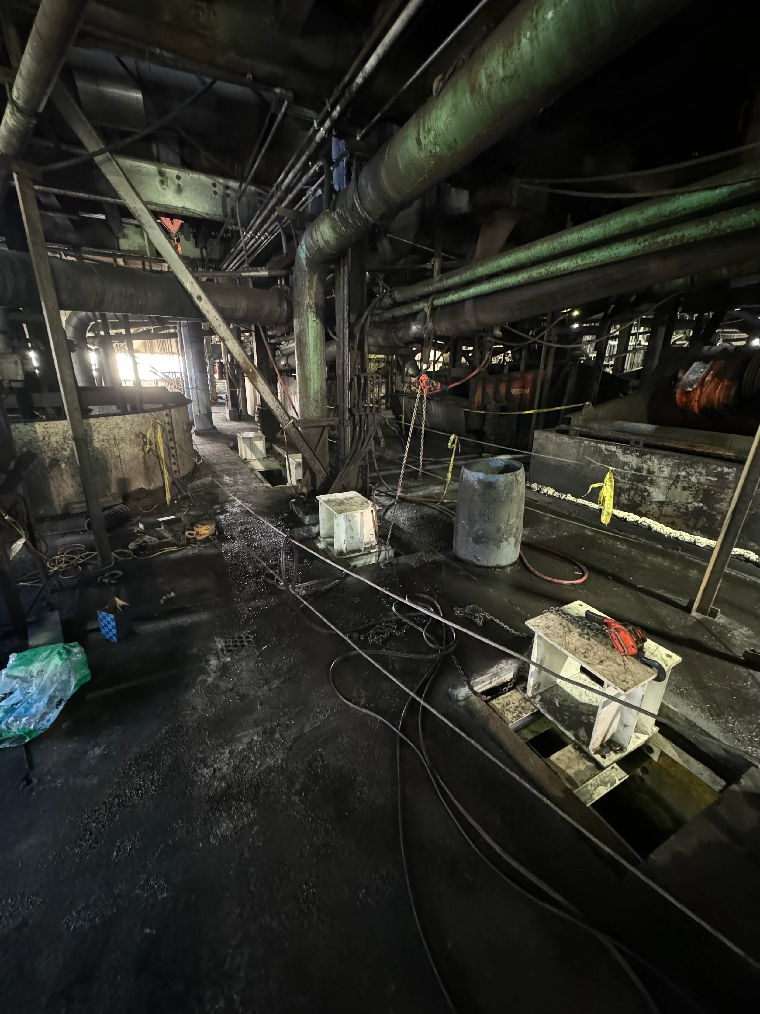Industrial interior with pipes, machinery, and equipment, some covered in grime and rust, with scattered tools and supplies on the floor.