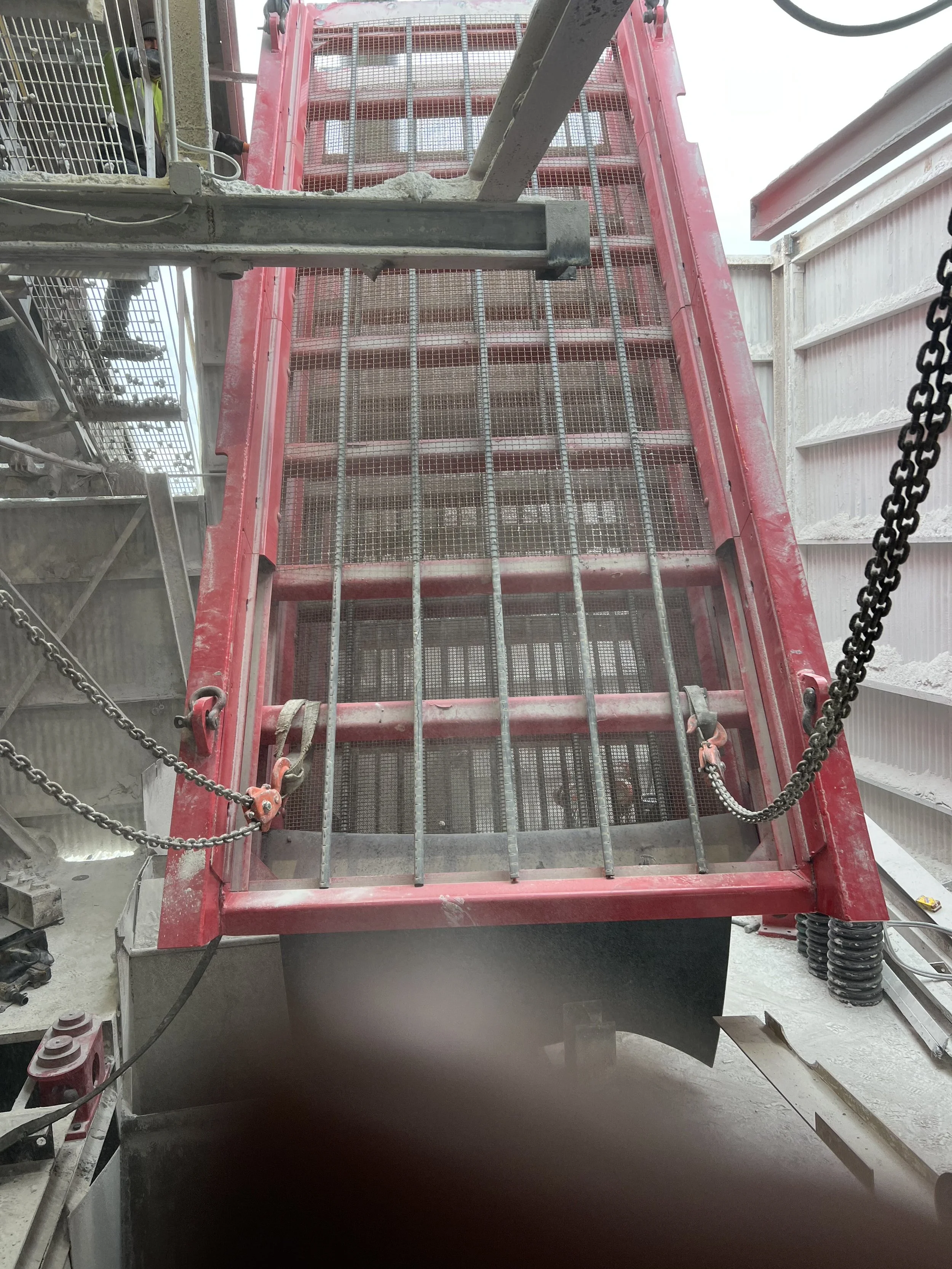 Inside view of a construction site with a large red metal structure, scaffolding, chains, and construction workers working on the site.