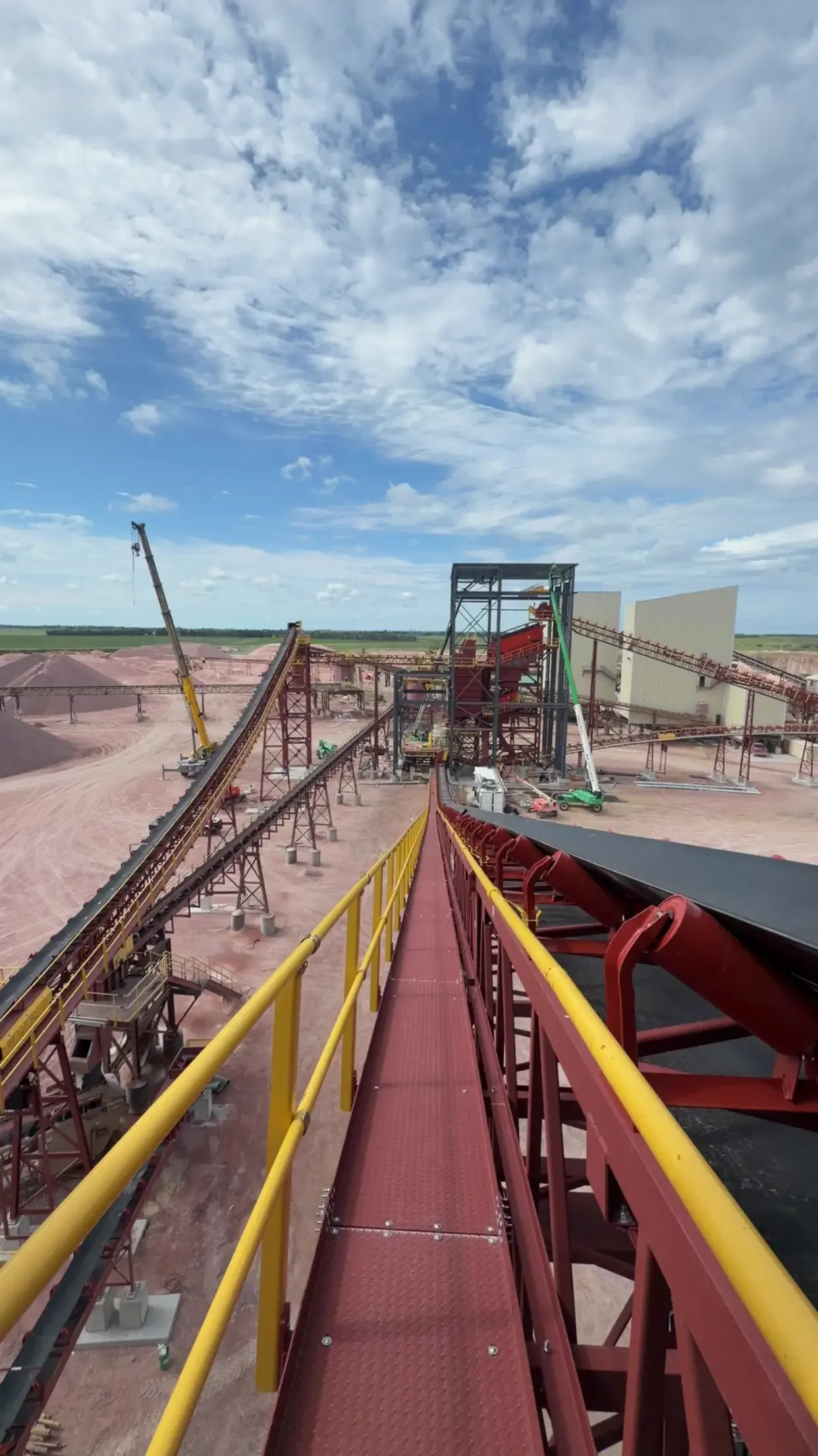 Construction site with red conveyor belts, cranes, and scaffolding under a blue sky with clouds.
