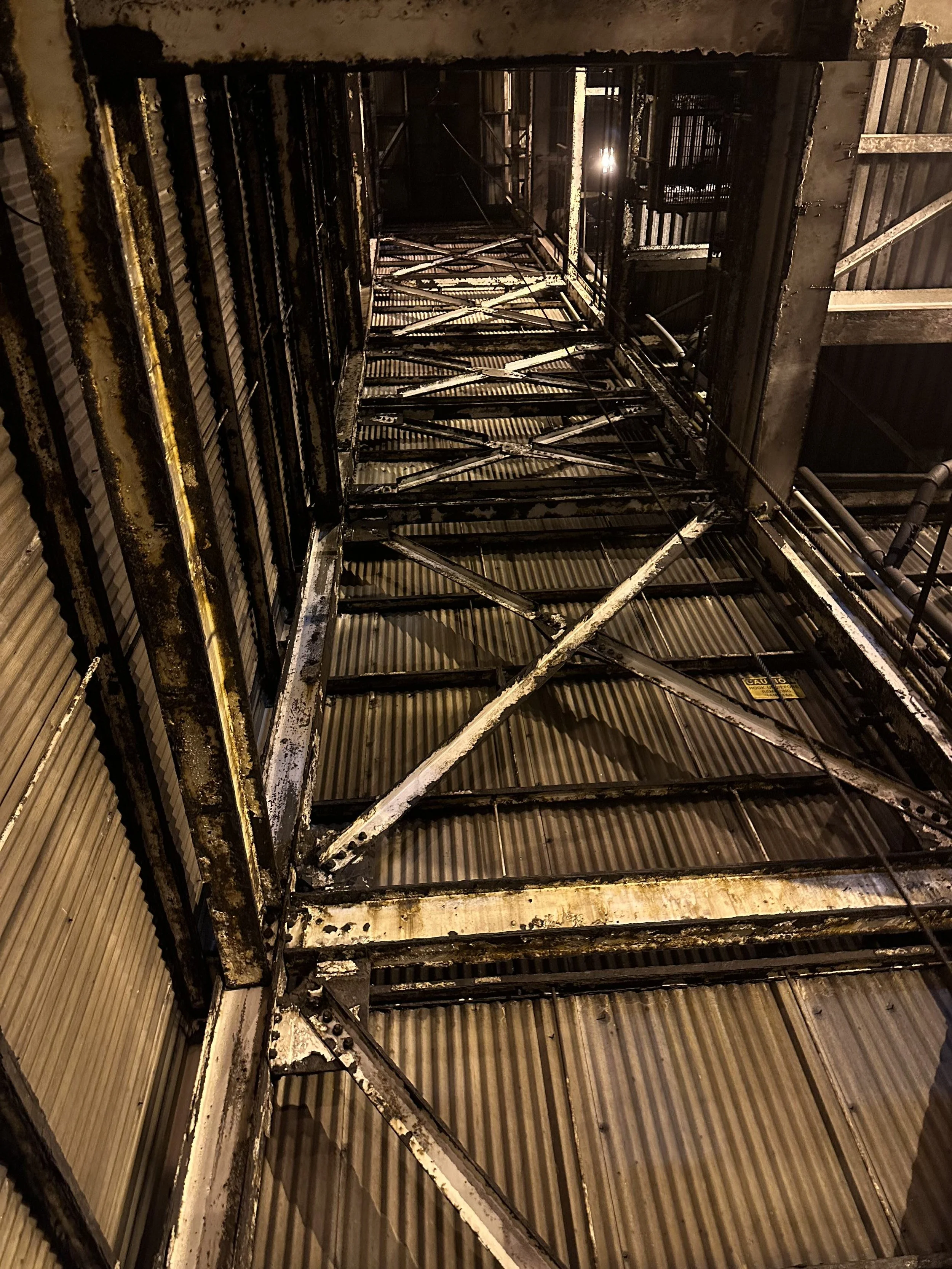Looking up at a deteriorated metal staircase with rust and peeling paint, inside an industrial building with corrugated metal walls.