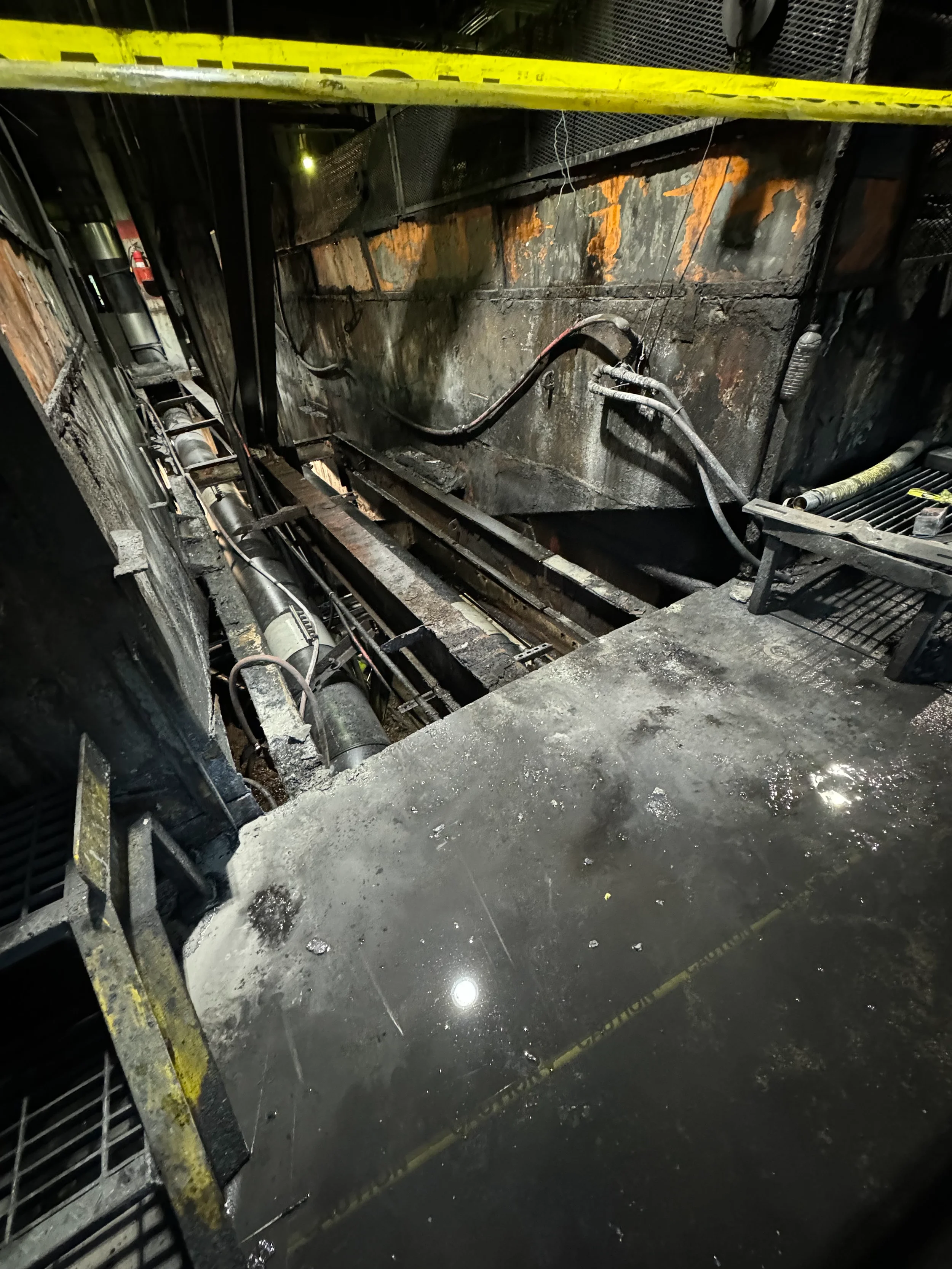 Burned and charred wall with scattered debris and water on the floor, located behind yellow caution tape in an industrial or construction setting.
