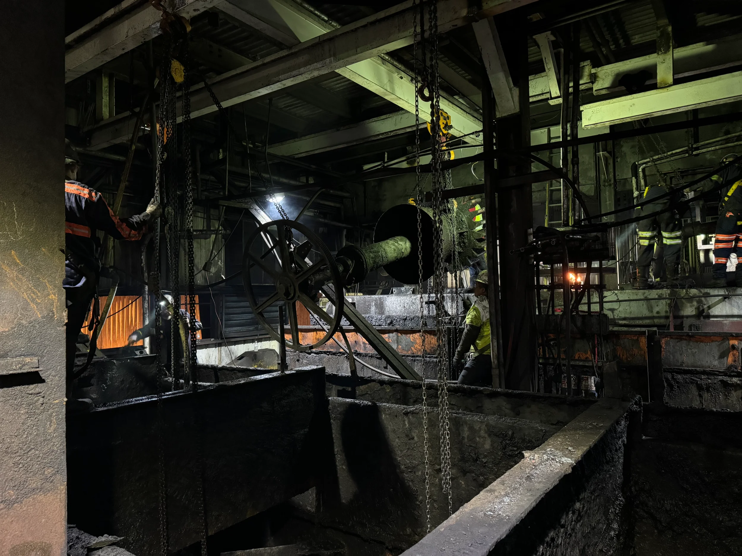 Workers in protective gear working inside an industrial facility with heavy machinery, chains, and large pipes, illuminated by industrial lighting.