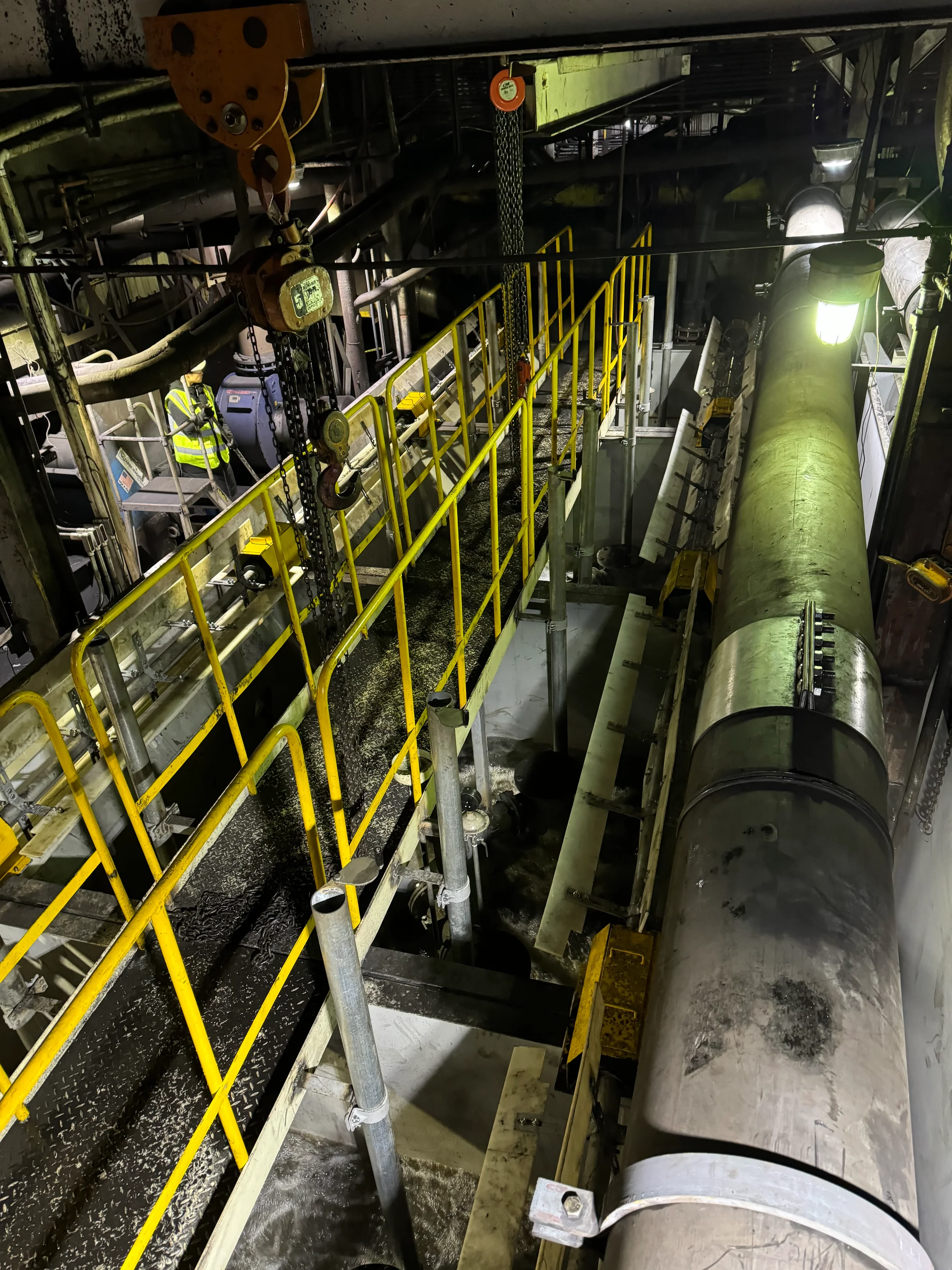 Industrial infrastructure with pipes, yellow safety railings, a large metal cylinder, a worker wearing a safety vest, and machinery in a dark industrial setting.