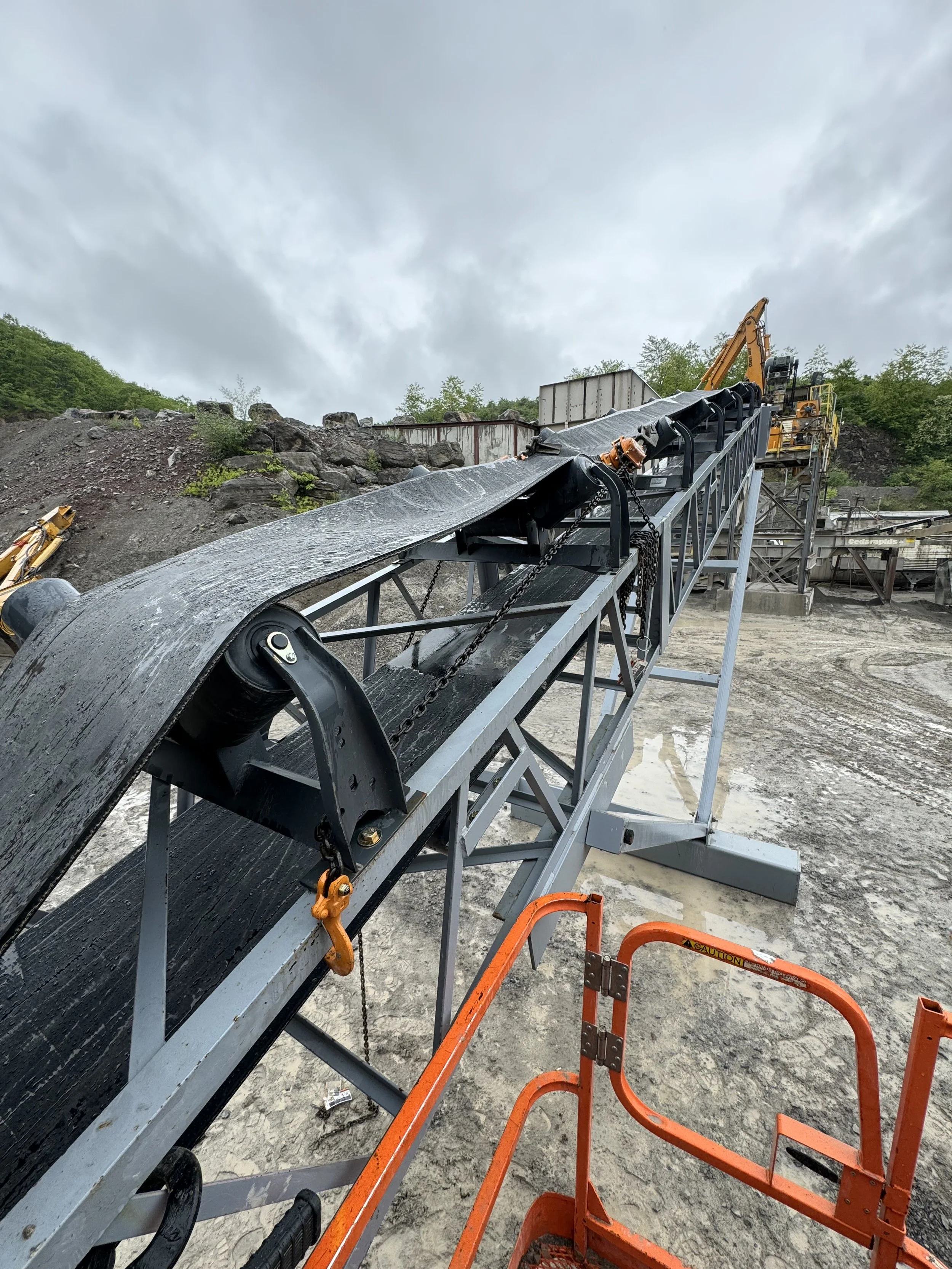 A conveyor belt system at a quarry or construction site, with a black rubber conveyor belt and an orange safety barrier in the foreground, set against a cloudy sky and rocky hillside.