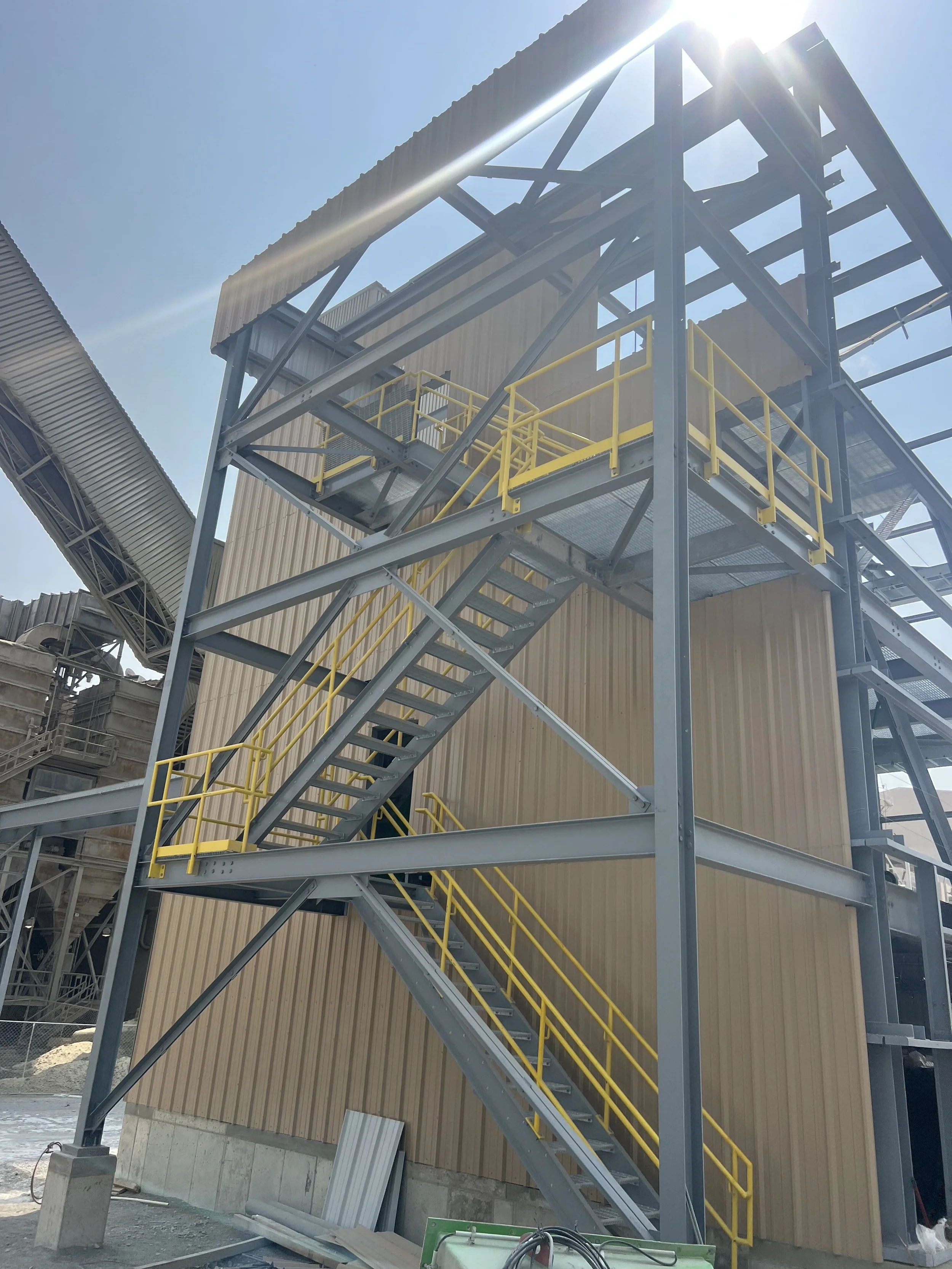 Construction site with a metal staircase with yellow railings leading to a building under construction with beige walls and a corrugated metal roof, during a sunny day.