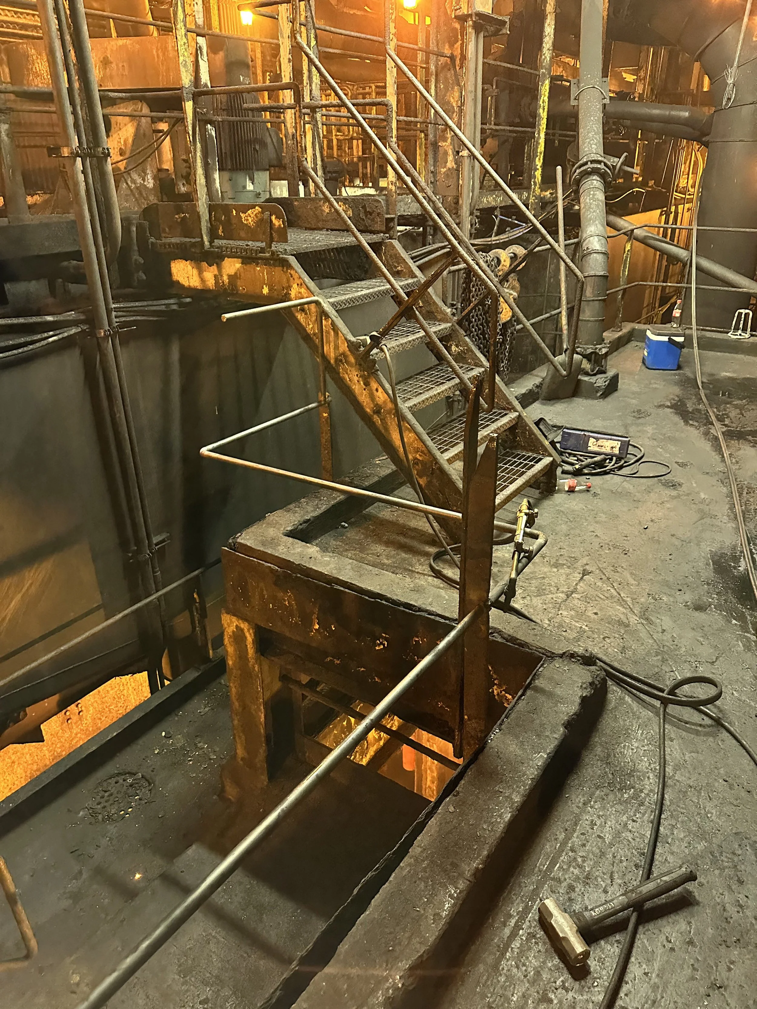 Industrial scene with rusted stairs, pipes, and machinery inside a factory or metalworks, illuminated by orange lighting.