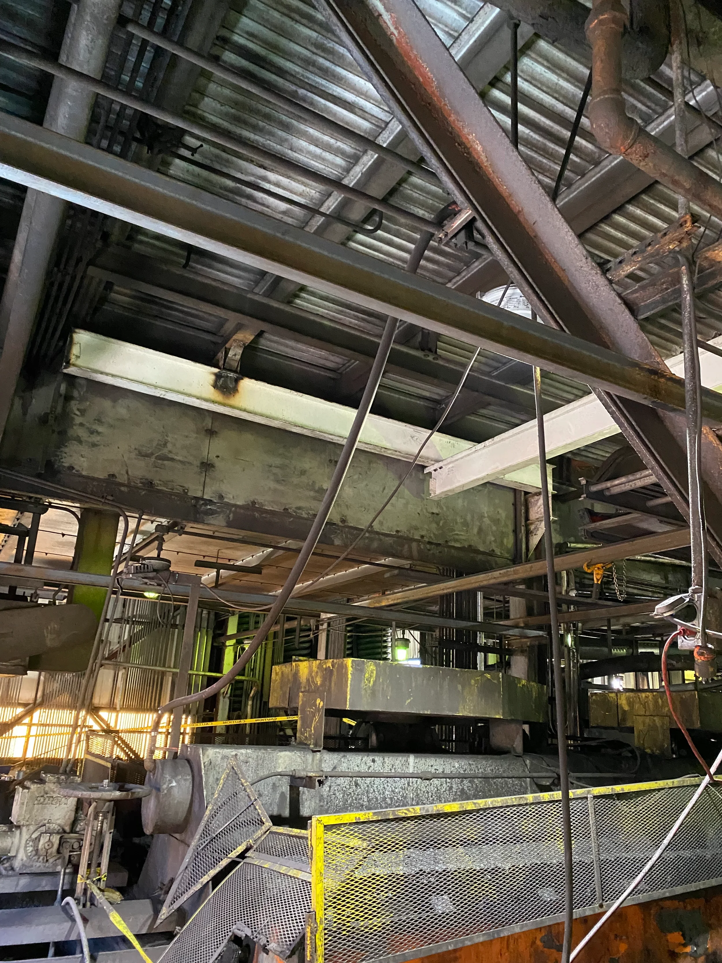 Industrial setting with machinery, metal piping, and safety barriers, illuminated with dim lighting.