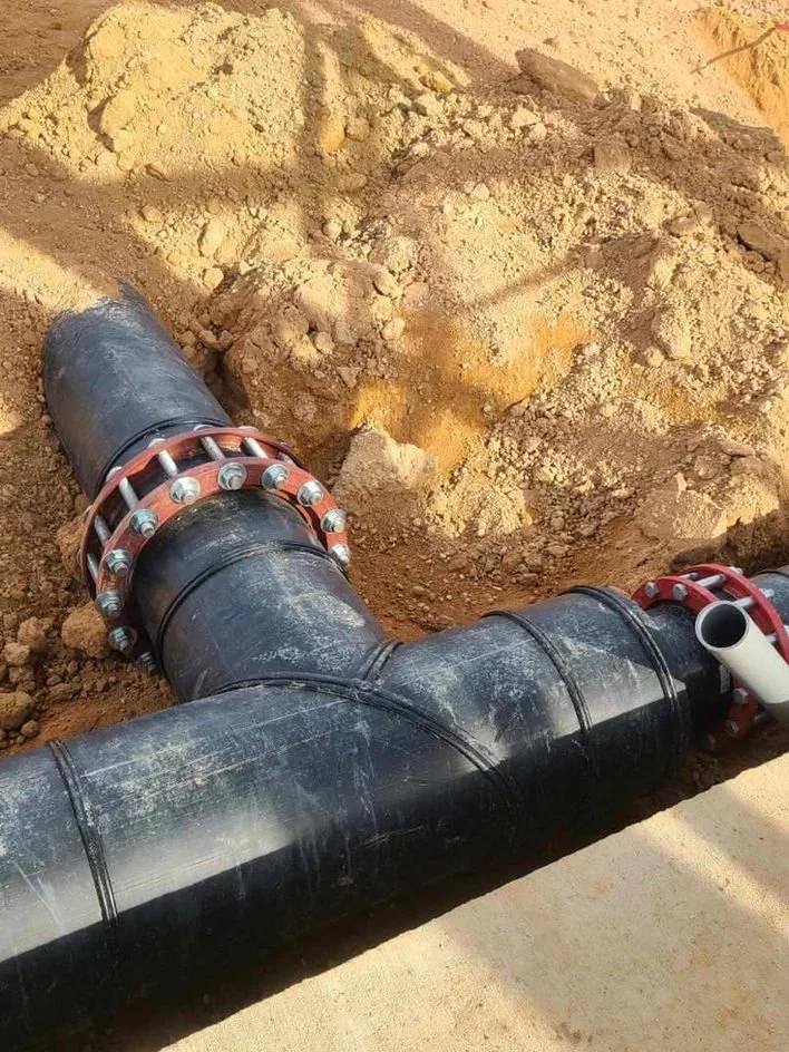 Close-up of large black industrial pipes with red and silver couplings, lying on dry dirt at a construction site.