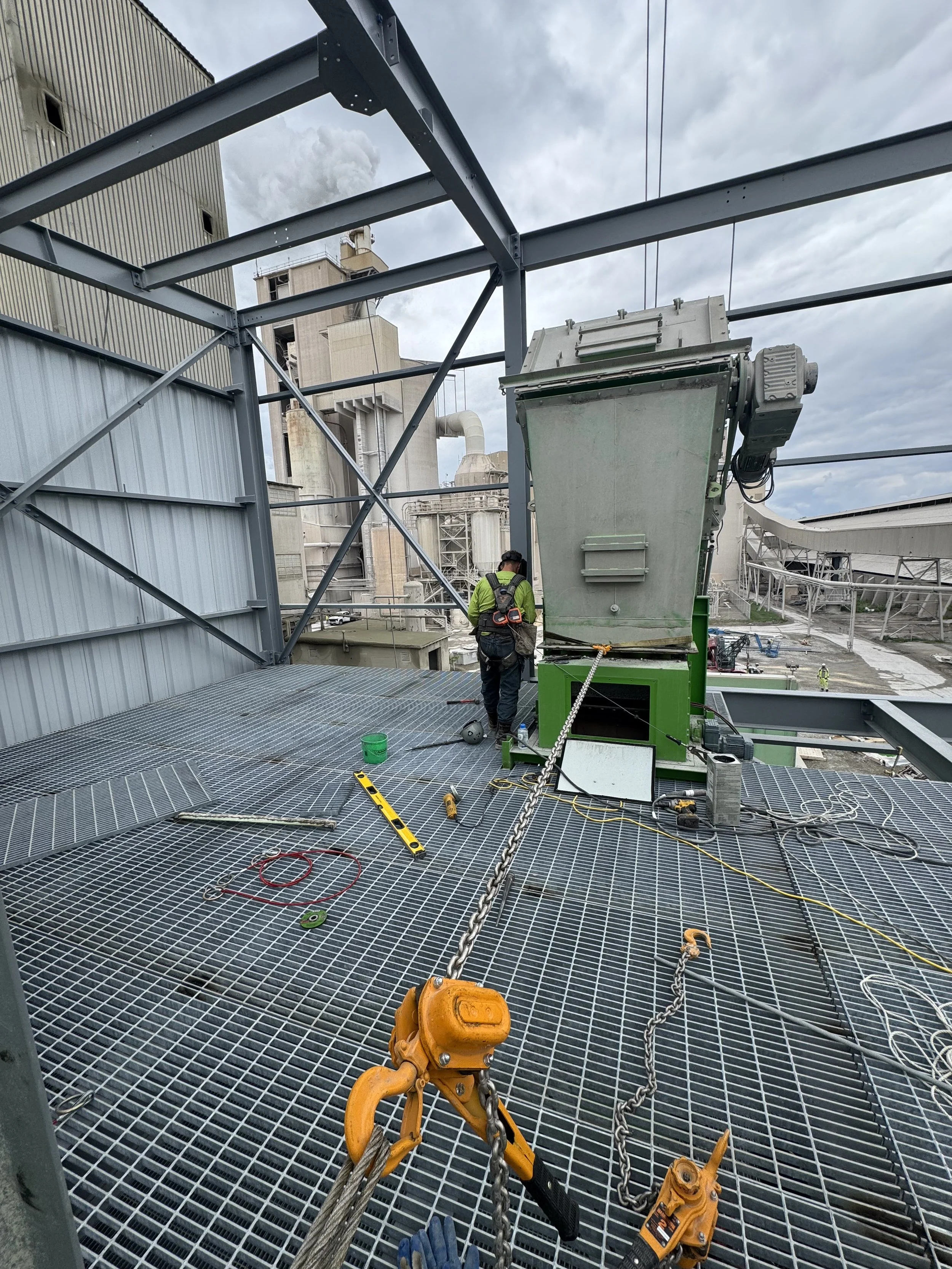 Construction worker on metal grated platform installing large industrial machinery, with factory buildings and cloudy sky in the background.