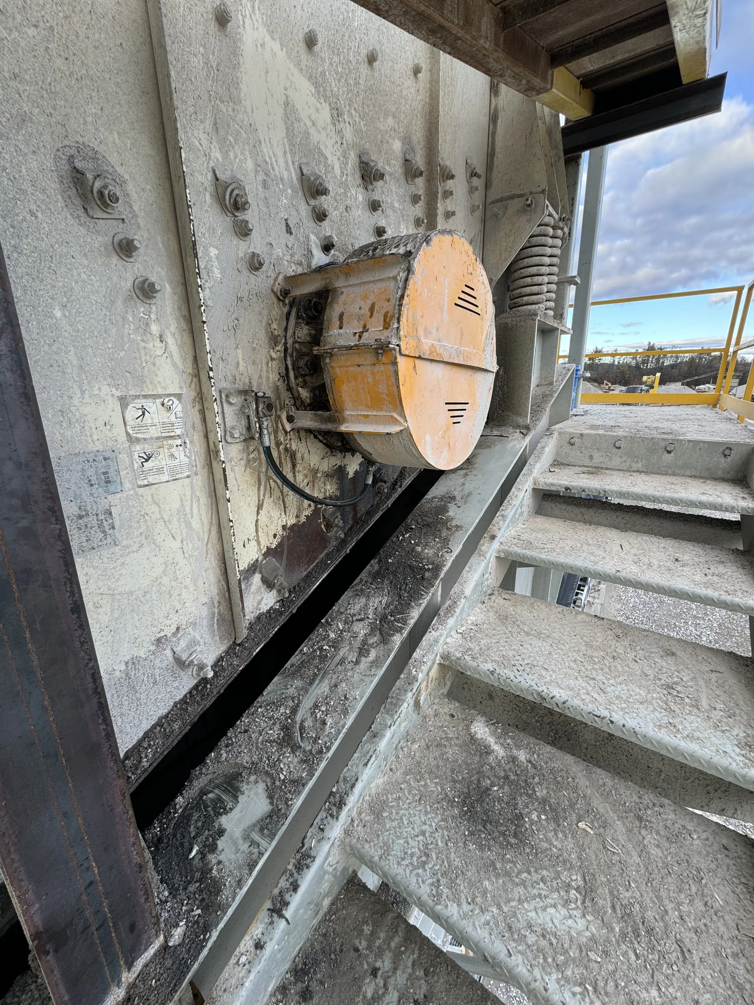 Close-up of a construction site showing construction equipment, metal platform, bolts, a large orange motor or gear in the foreground, and a backdrop of construction materials and partly cloudy sky.