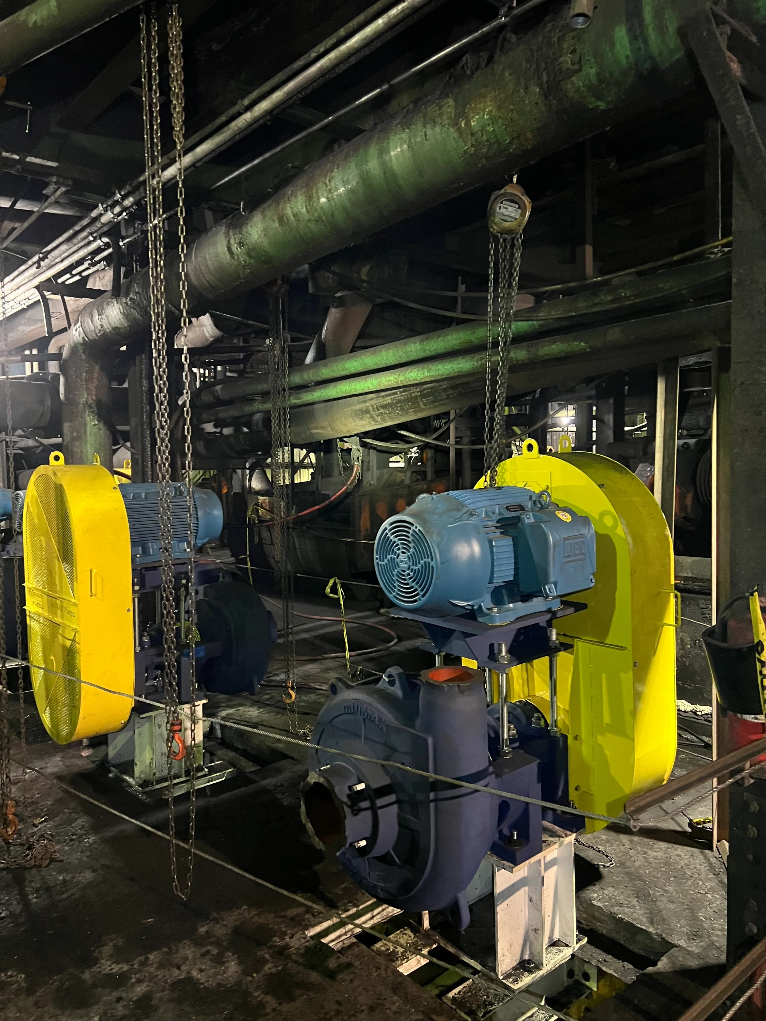 Industrial machinery with large blue electric motors, yellow safety covers, chains, and pipes in a dark factory setting.