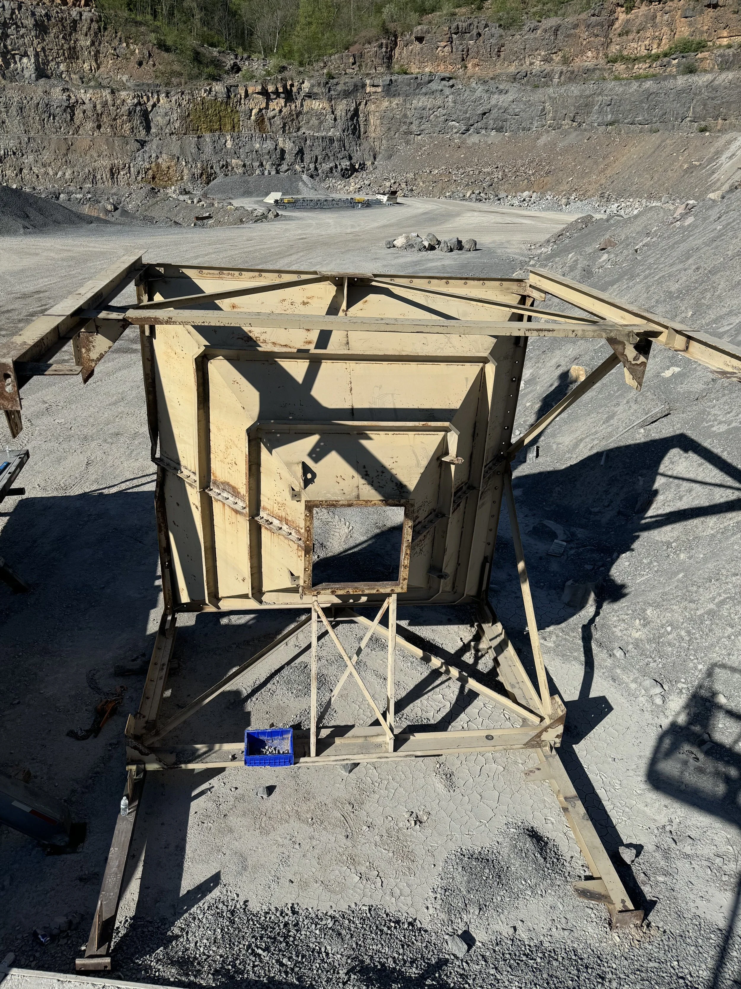 View of a gravel quarry with machinery and a large metal frame in the foreground, and a cliff with rocky layers in the background, during outdoor daylight.