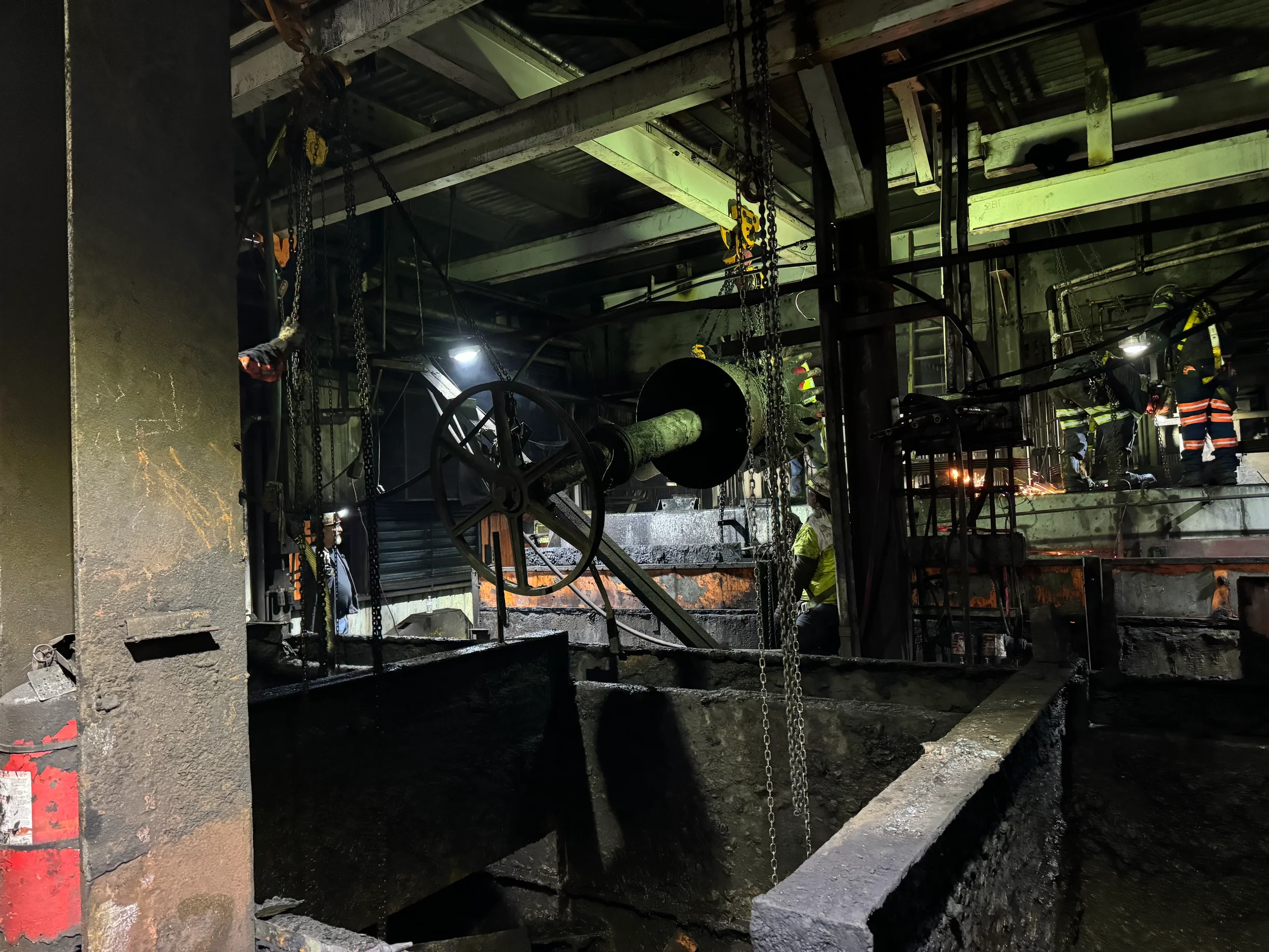 Construction workers welding inside an industrial building with pipes and chains hanging from the ceiling at night.