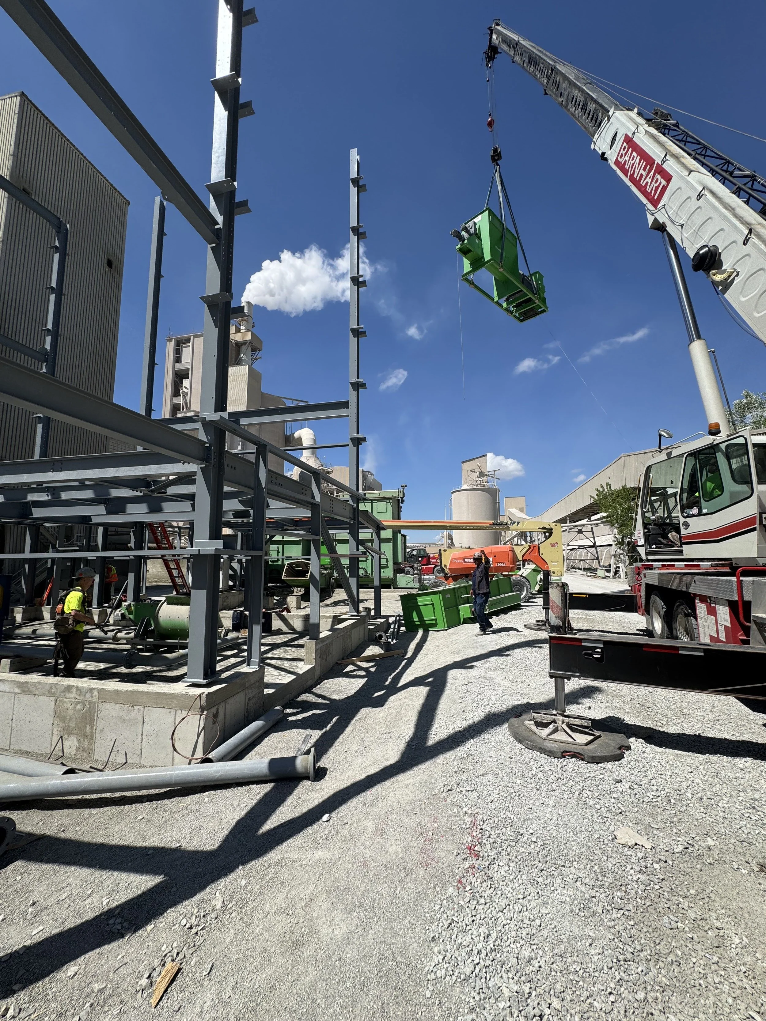 Construction site with steel framework, a crane lifting green equipment, workers, and partially built structures under a blue sky.