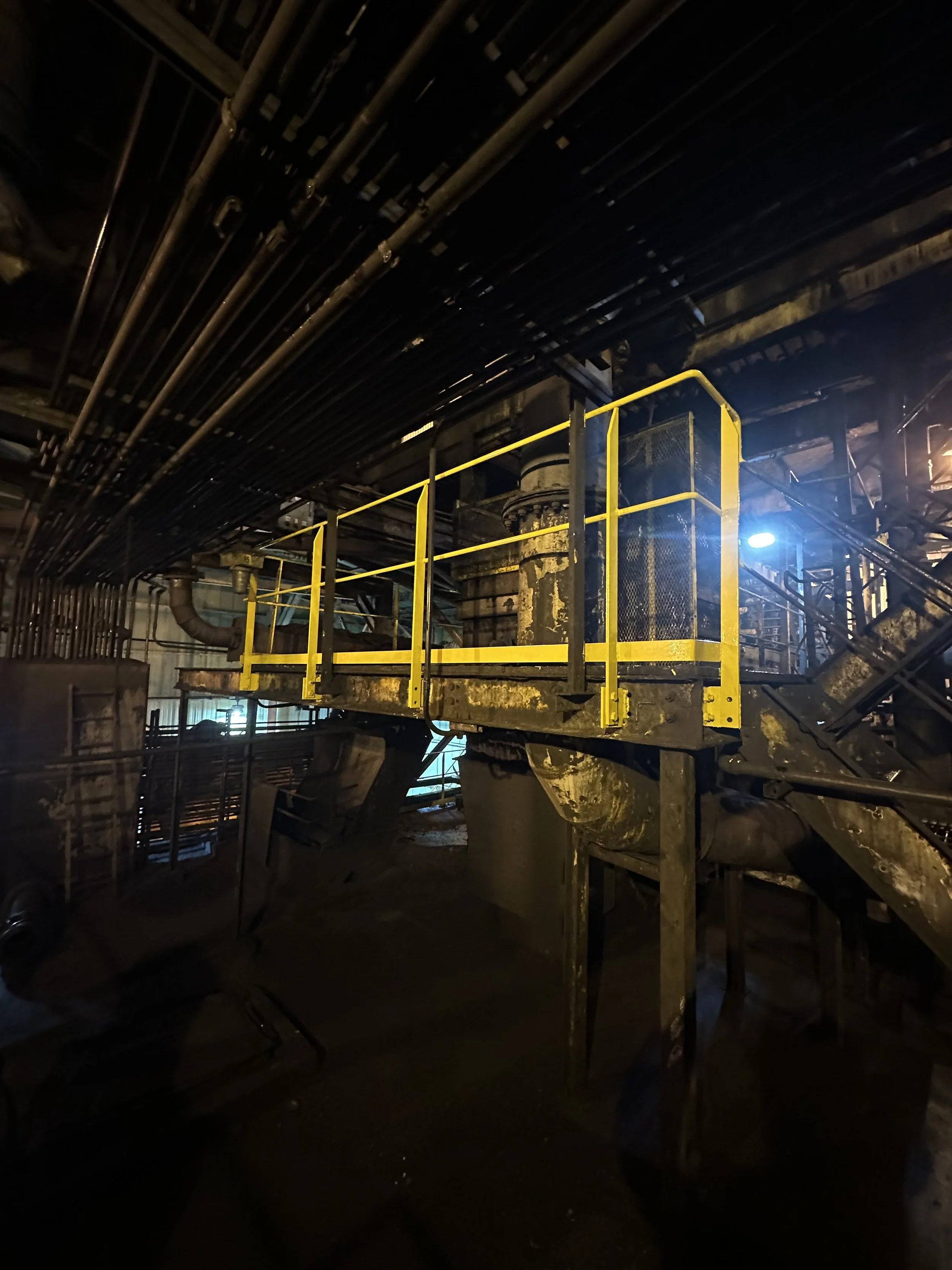Industrial underground setting with pipes overhead and a yellow safety railing around a platform, dimly lit with a blue light