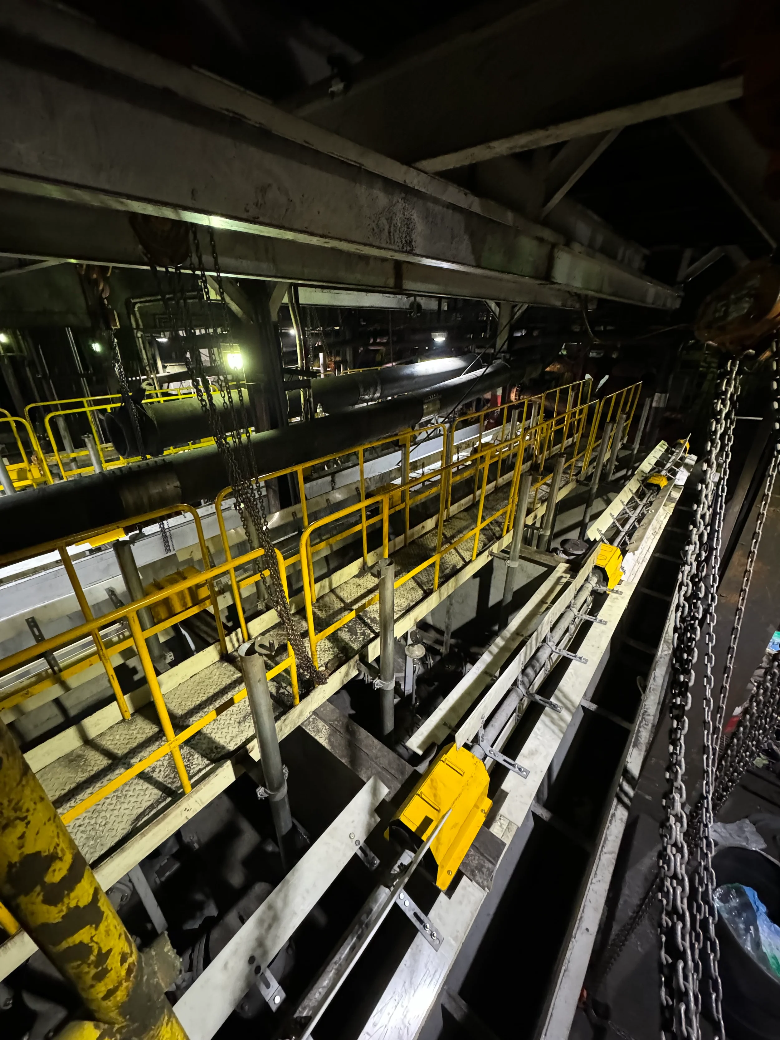 Interior view of an industrial or factory setting with metal pipes, yellow safety railings, chains, and machinery in a dark environment