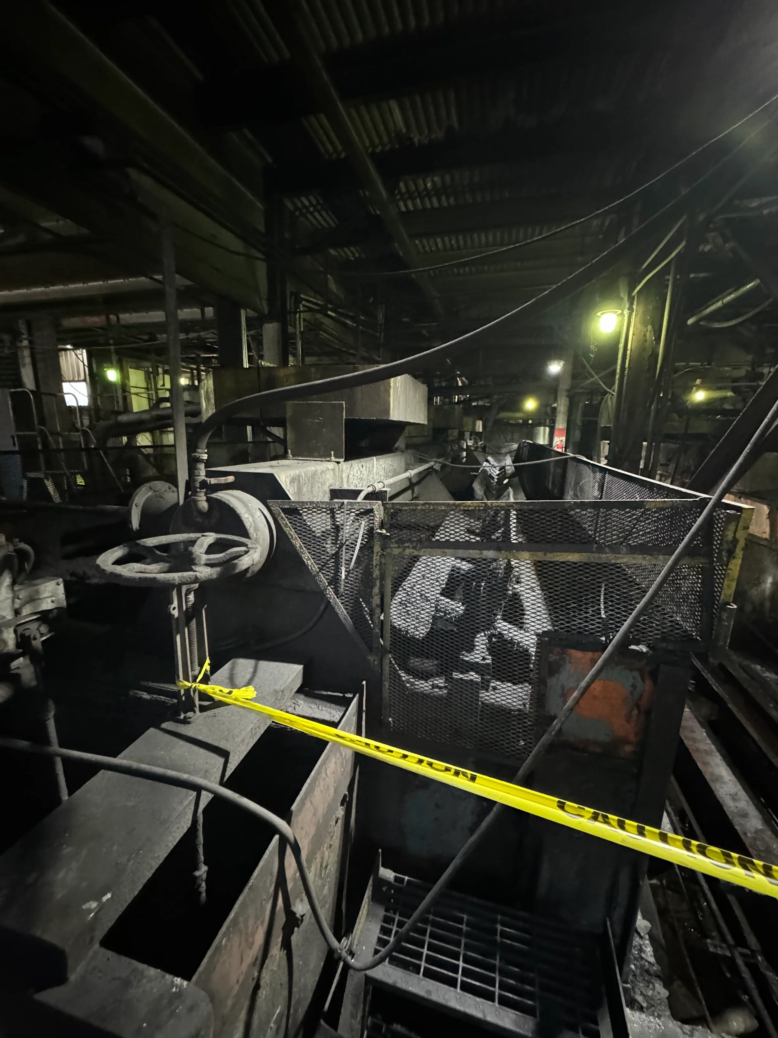 Dark industrial interior with metal stairs and walkways, yellow caution tape, and dim lighting from overhead fixtures.