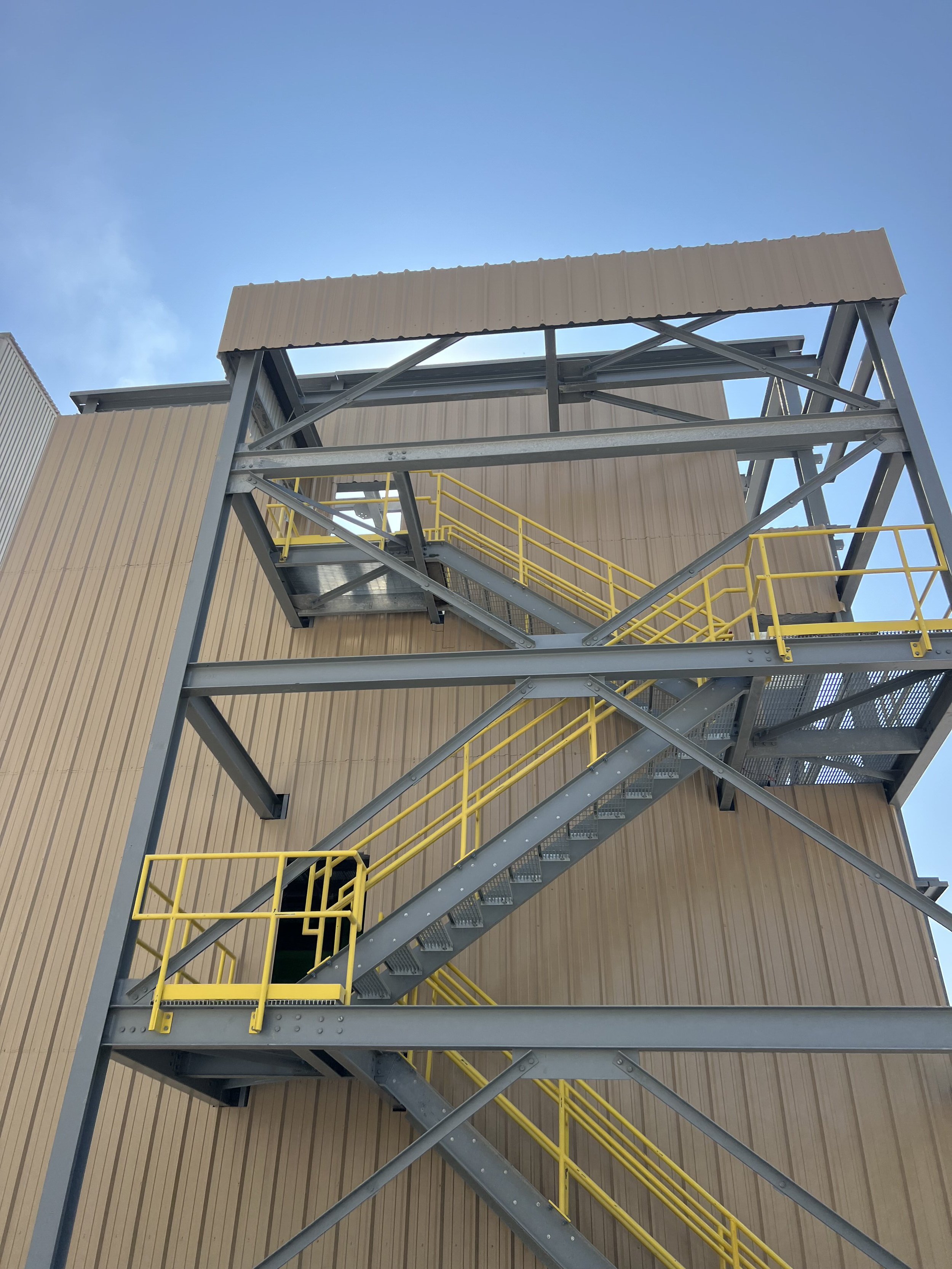 An outdoor metal staircase with yellow railings attached to a building with beige vertical siding, with a clear blue sky in the background.