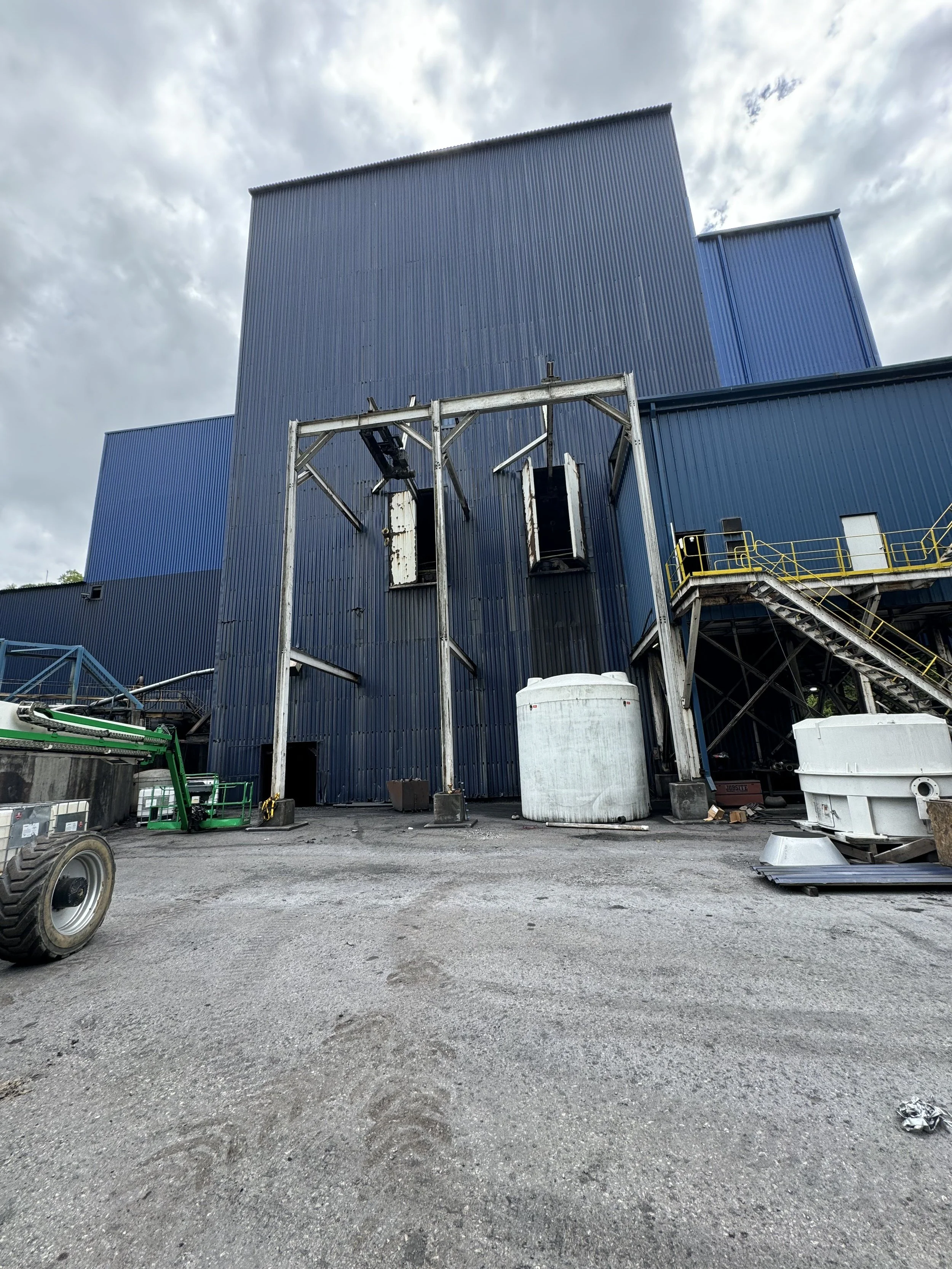 Industrial building with blue corrugated metal walls, stairs, and equipment outside, under cloudy sky.