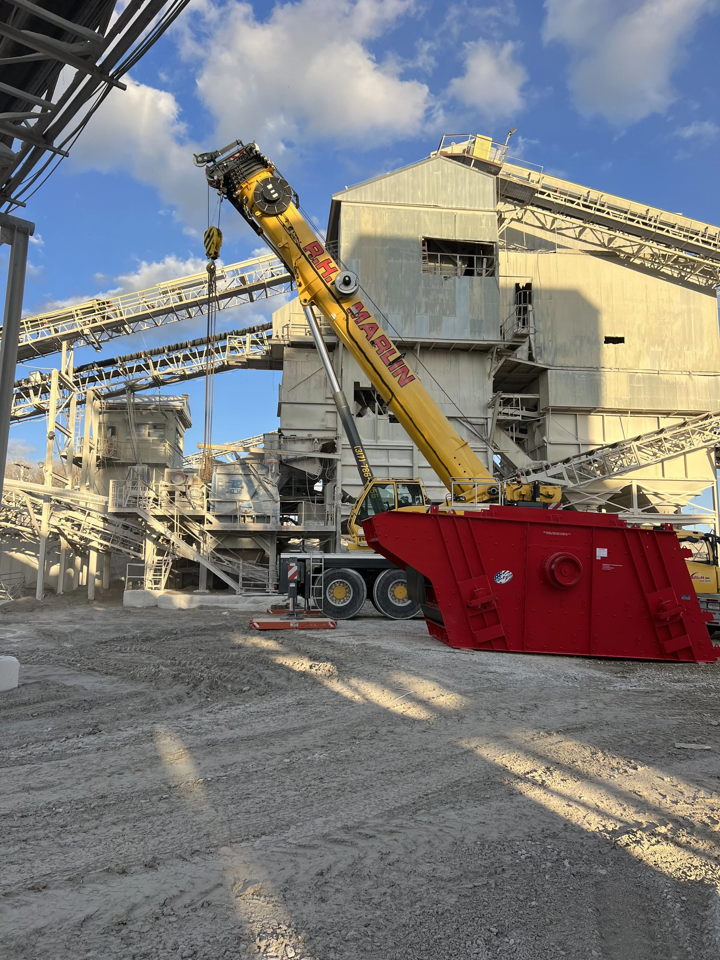 A construction site with a large yellow crane lifting equipment in front of a multi-story industrial building under a blue sky with clouds.