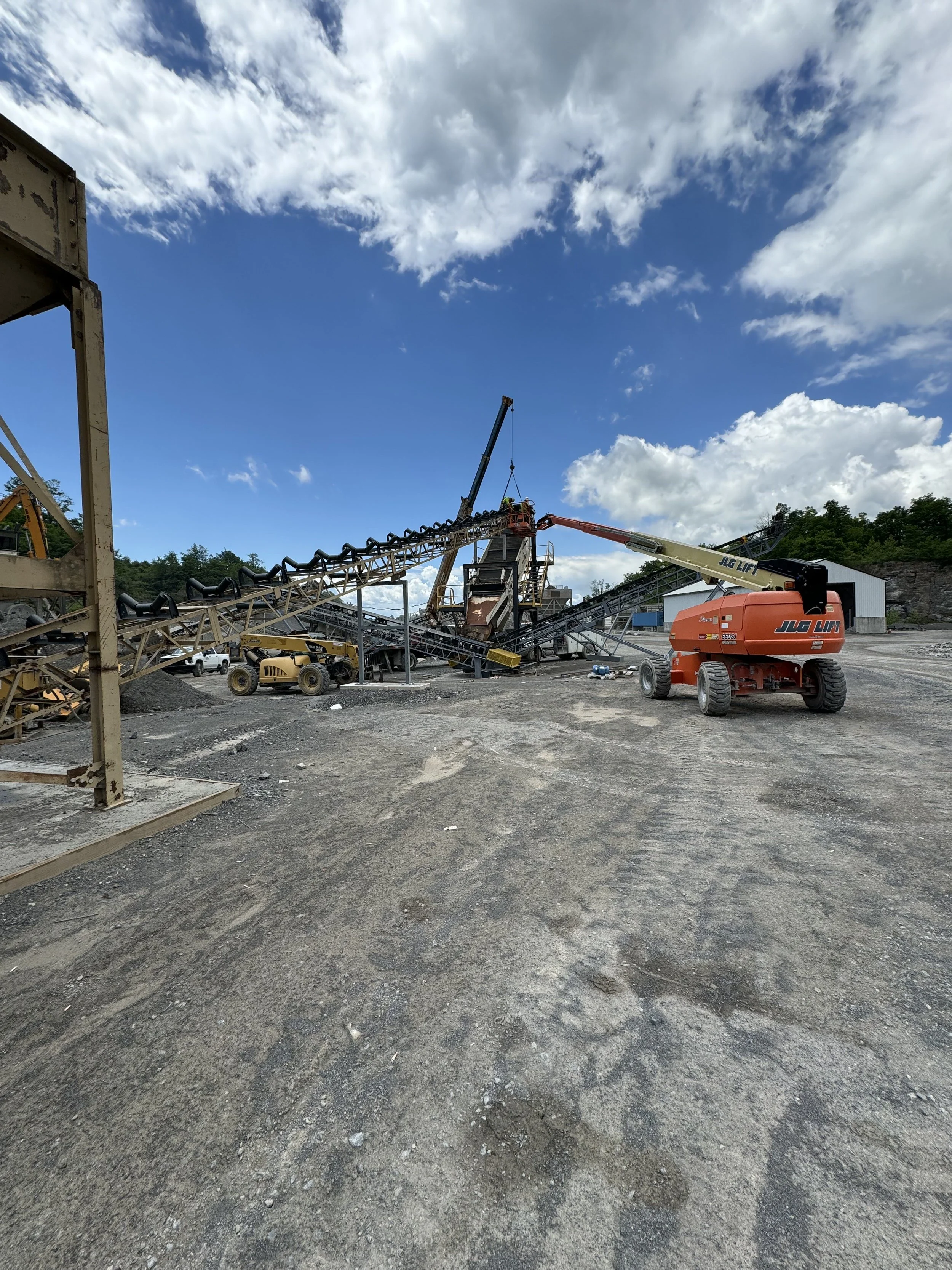 Construction site with heavy machinery, including a crane and an excavator, on a dirt ground under a partly cloudy sky.