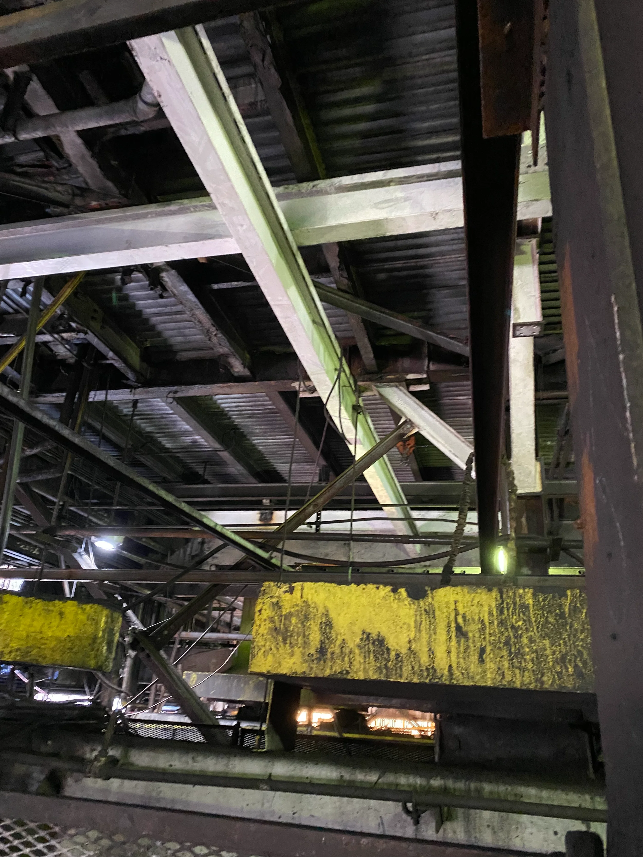 Close-up view of an industrial or construction site with metal beams, pipes, and grates, some of which are rusted or weathered, with yellow painted markings on a surface.
