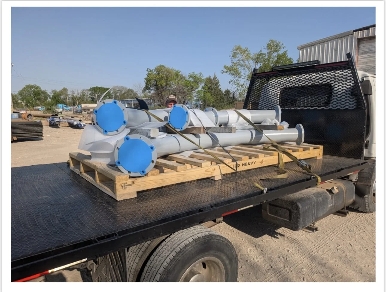 Flatbed truck carrying large white pipes with blue flanges, secured with straps on a wooden pallet