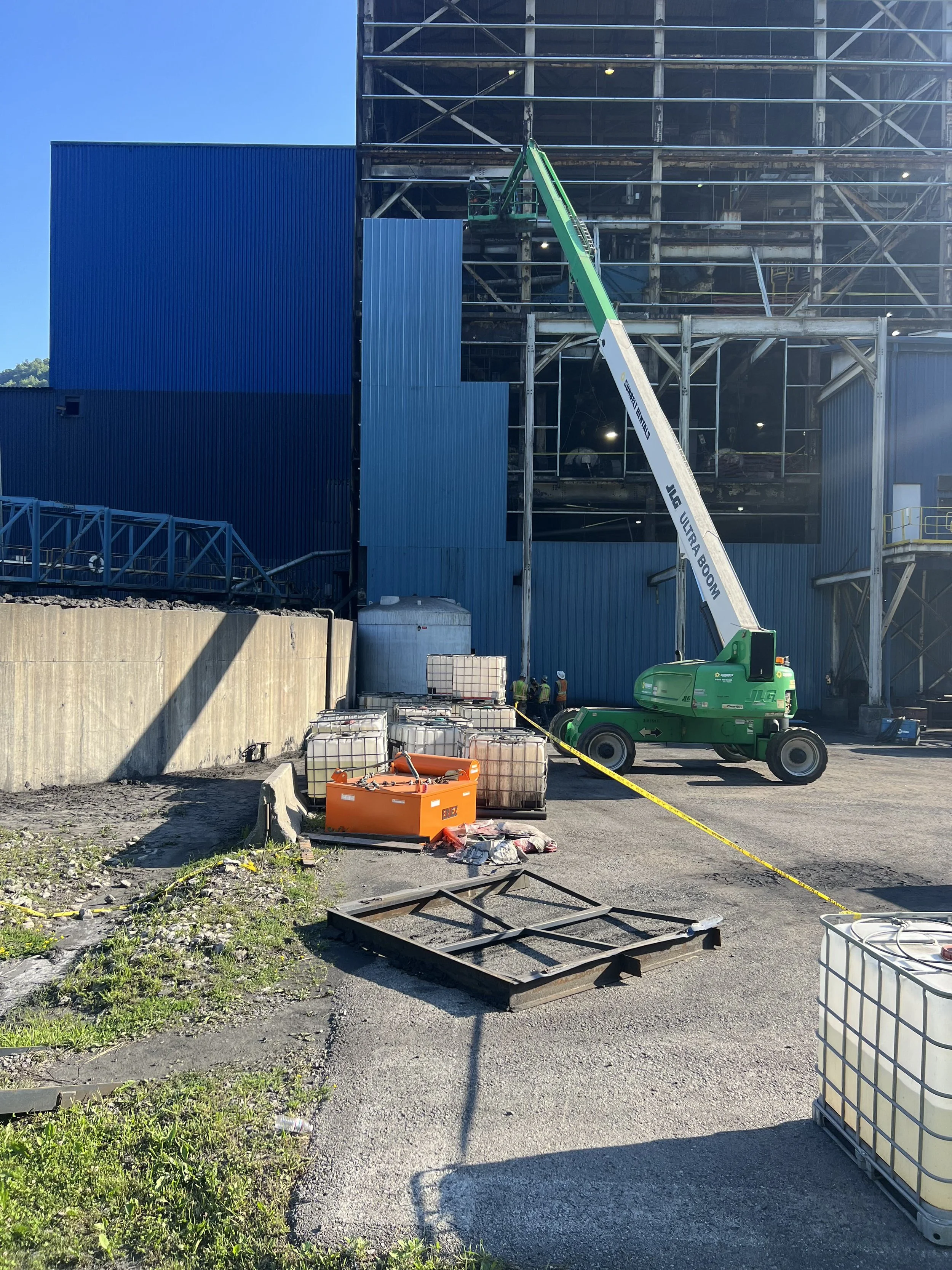 Construction site with a green telescopic boom lift, workers below, large building under construction with steel and glass framework, blue exterior wall, and various construction materials on the ground.