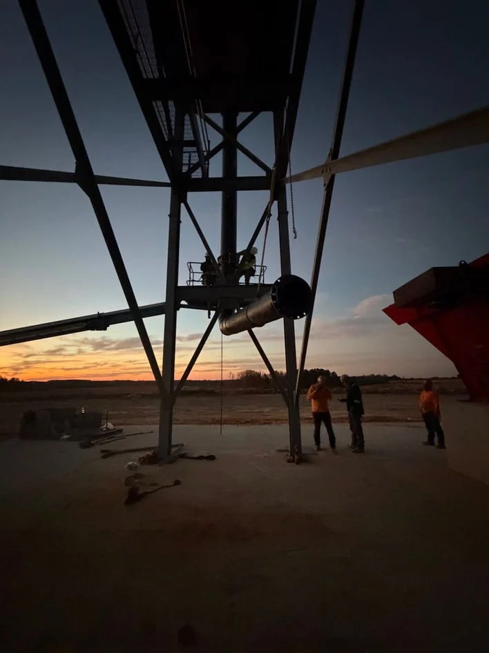 Workers inspecting a large satellite dish or radar structure during sunset, with a clear sky and open landscape in the background.