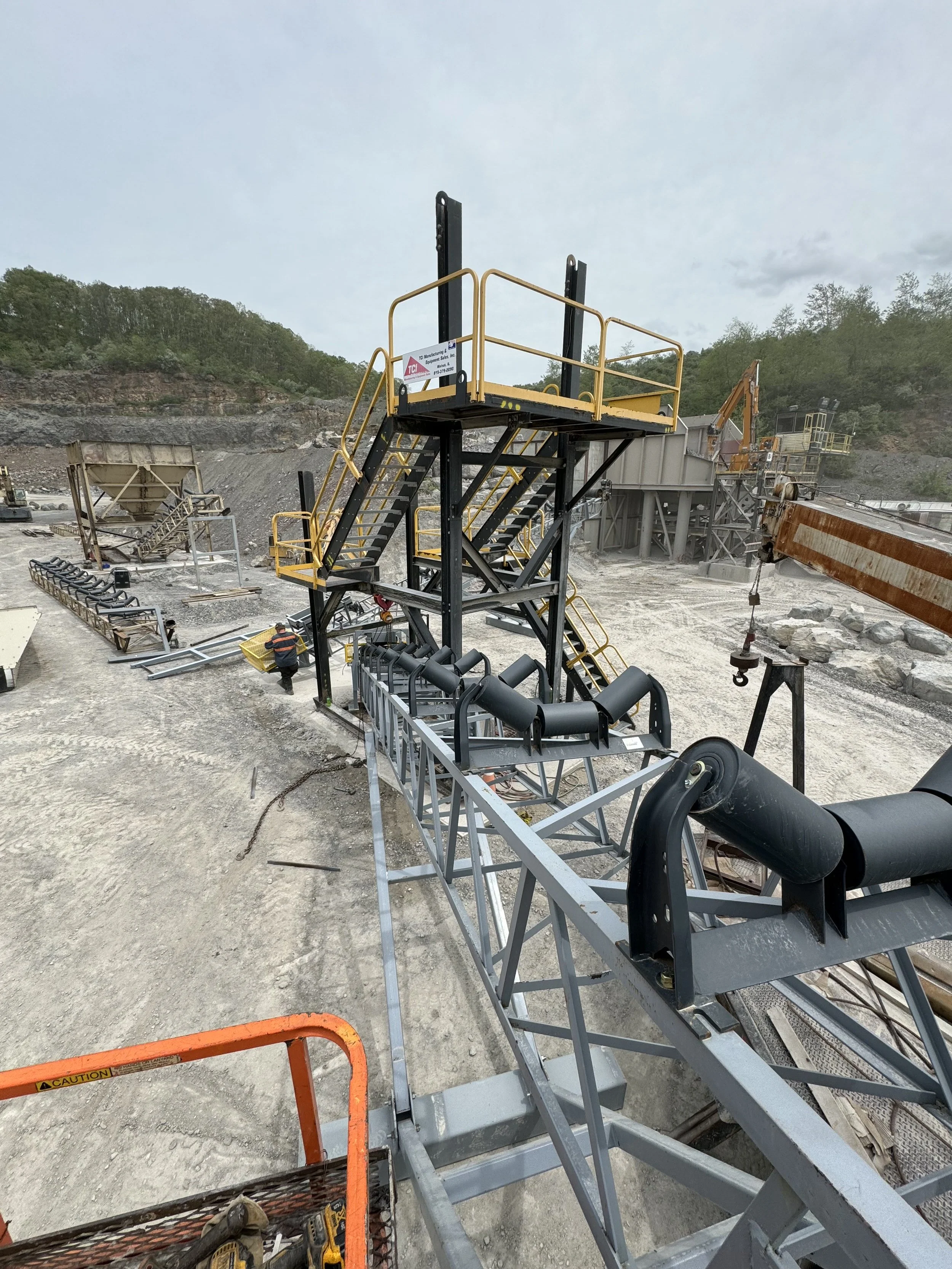 Construction site with conveyor belt system, stairs, and machinery in a rocky outdoor area, with hills and trees in the background.