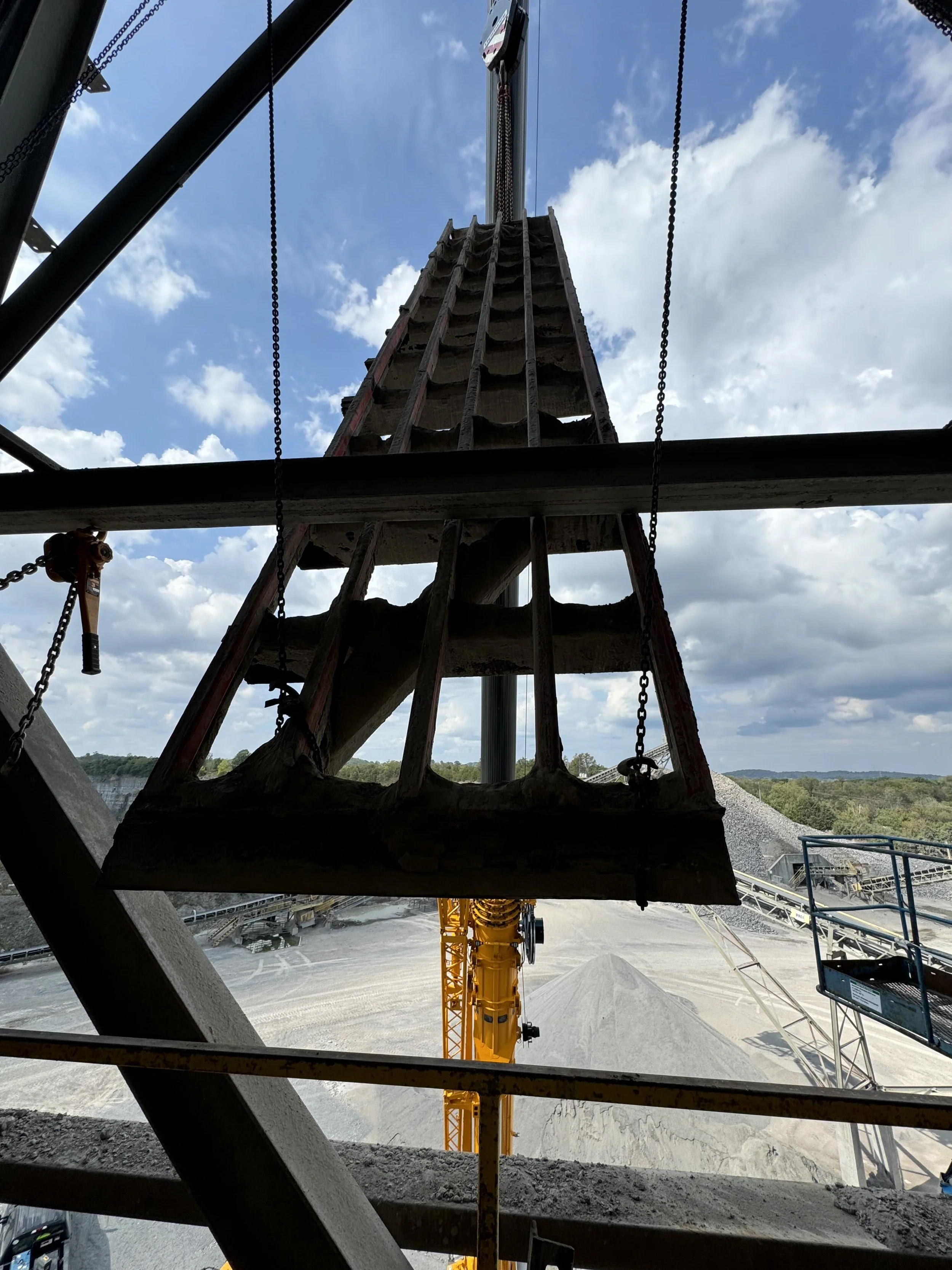 View from beneath a construction crane with a wooden platform, set against a blue sky with clouds, overlooking a construction site with gravel, equipment, and hills in the background.