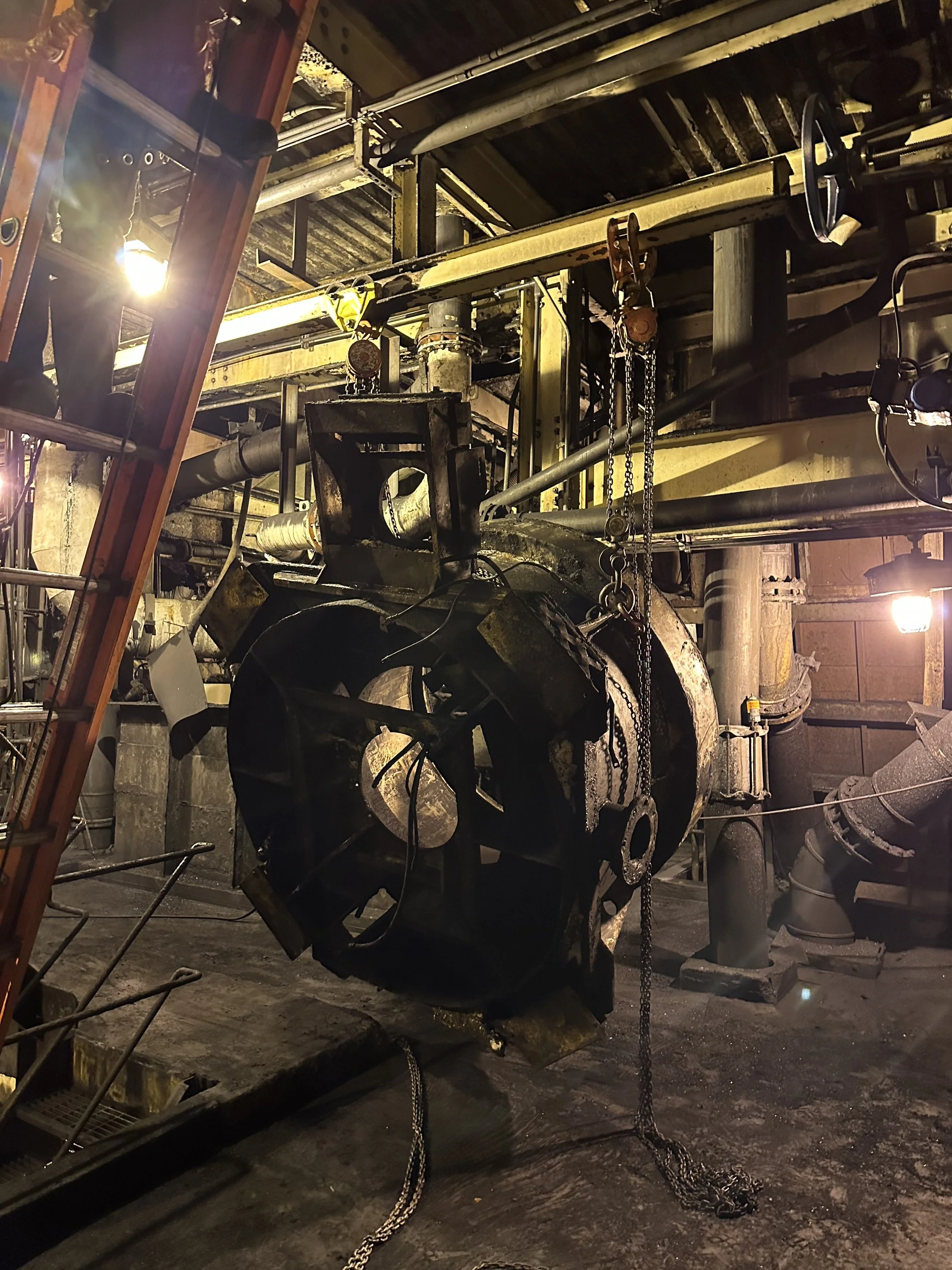 A close-up view of an industrial workshop with large machinery, chains, pipes, and metal structures. The area is illuminated with warm lighting, highlighting the rugged and worn surfaces of the equipment.