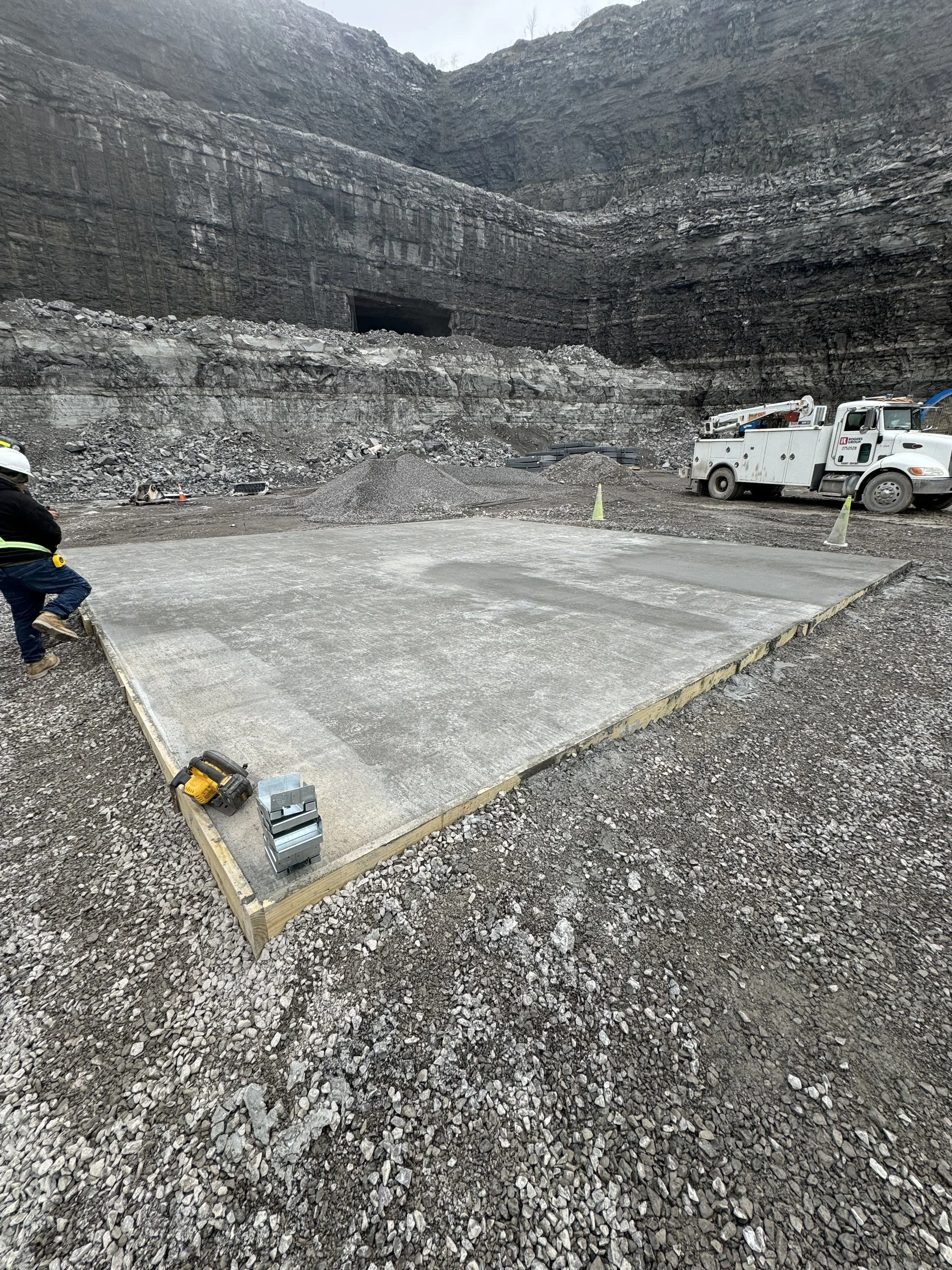 Construction workers are laying a concrete slab at a construction site surrounded by gravel and rock, with excavation walls in the background.
