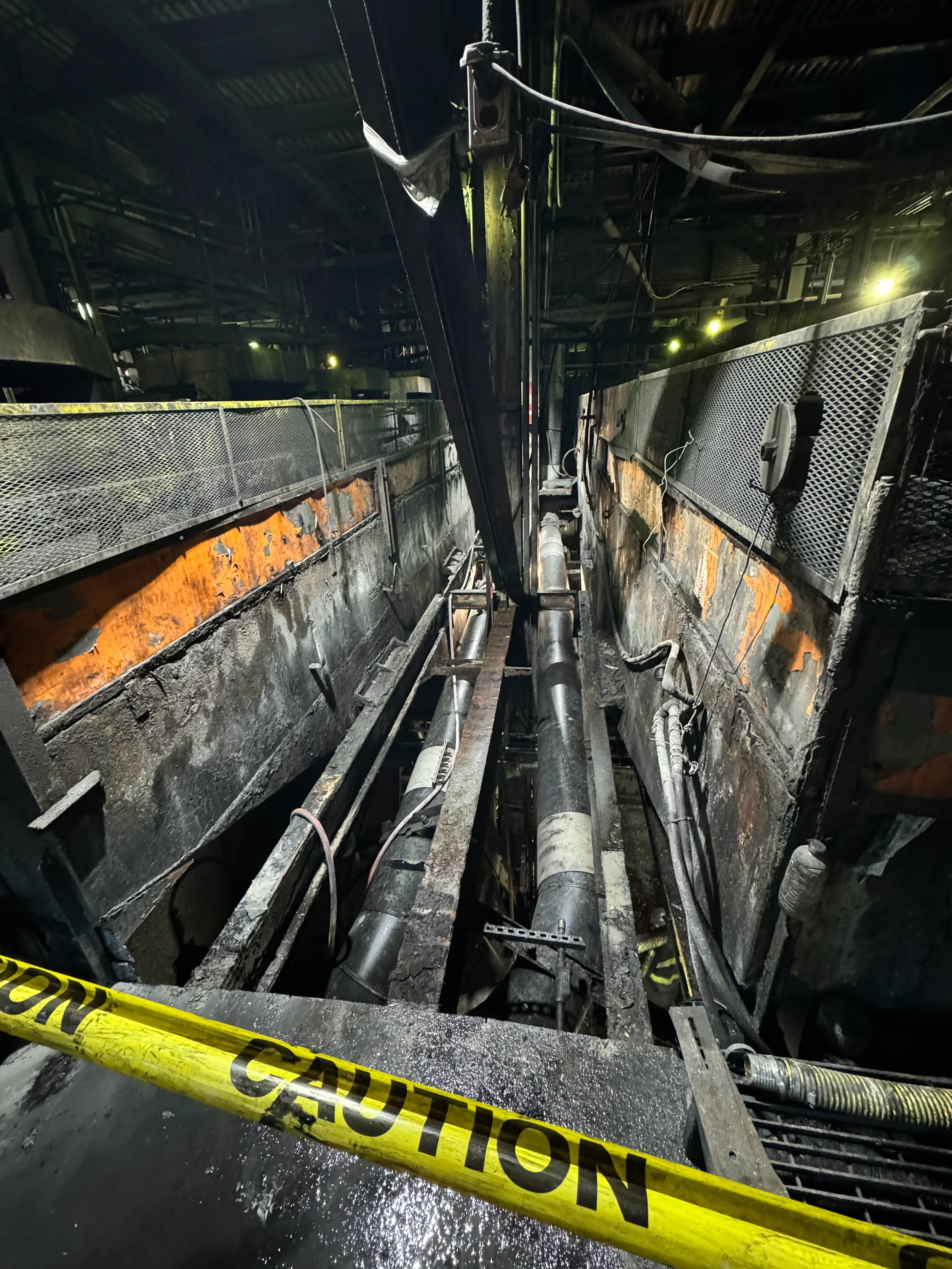 Industrial setting with damaged, charred metal structures and pipes, surrounded by yellow caution tape.
