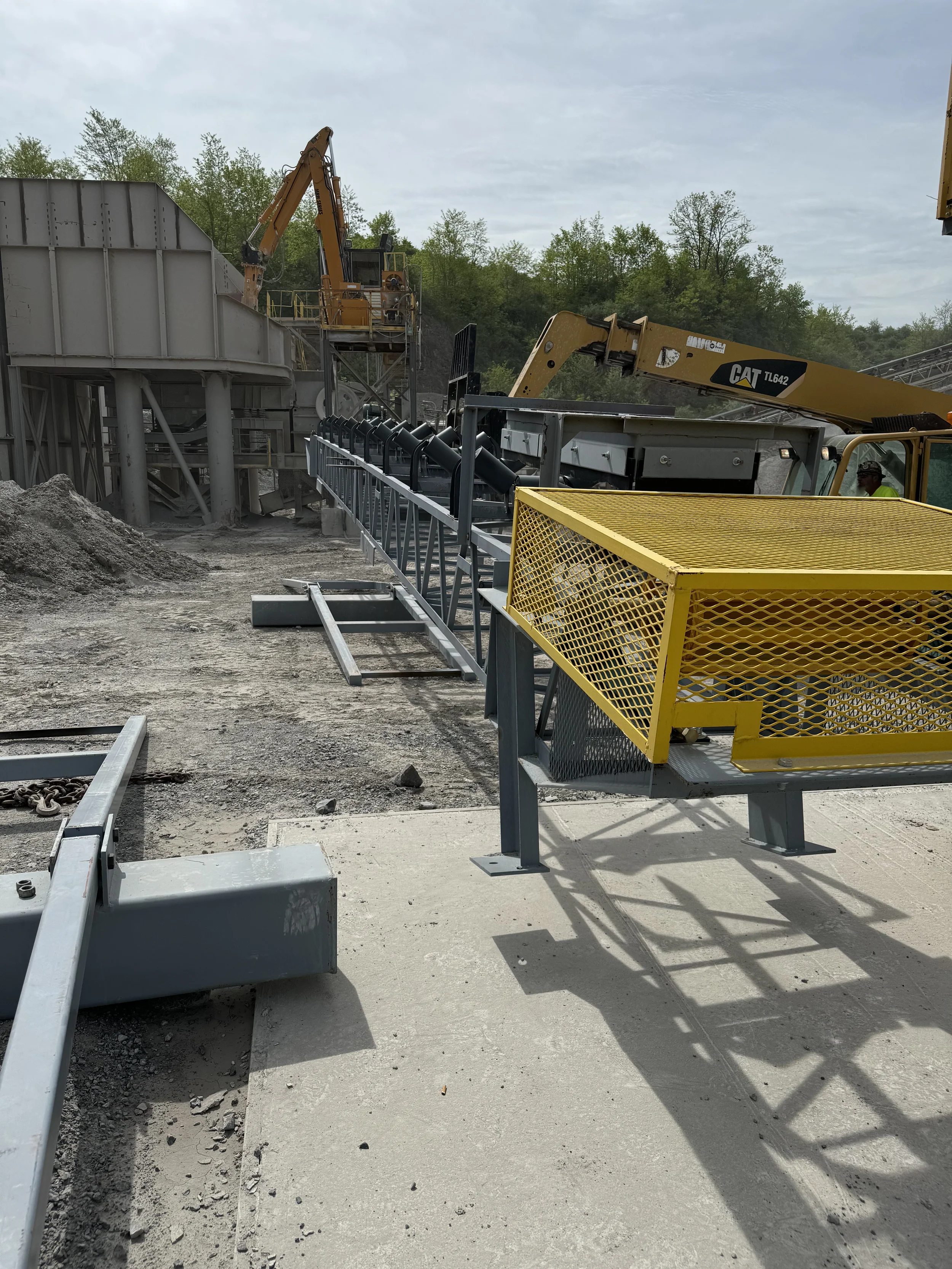 Construction site with yellow and gray scaffolding, machinery, and worker wearing a helmet, surrounded by trees and overcast sky.