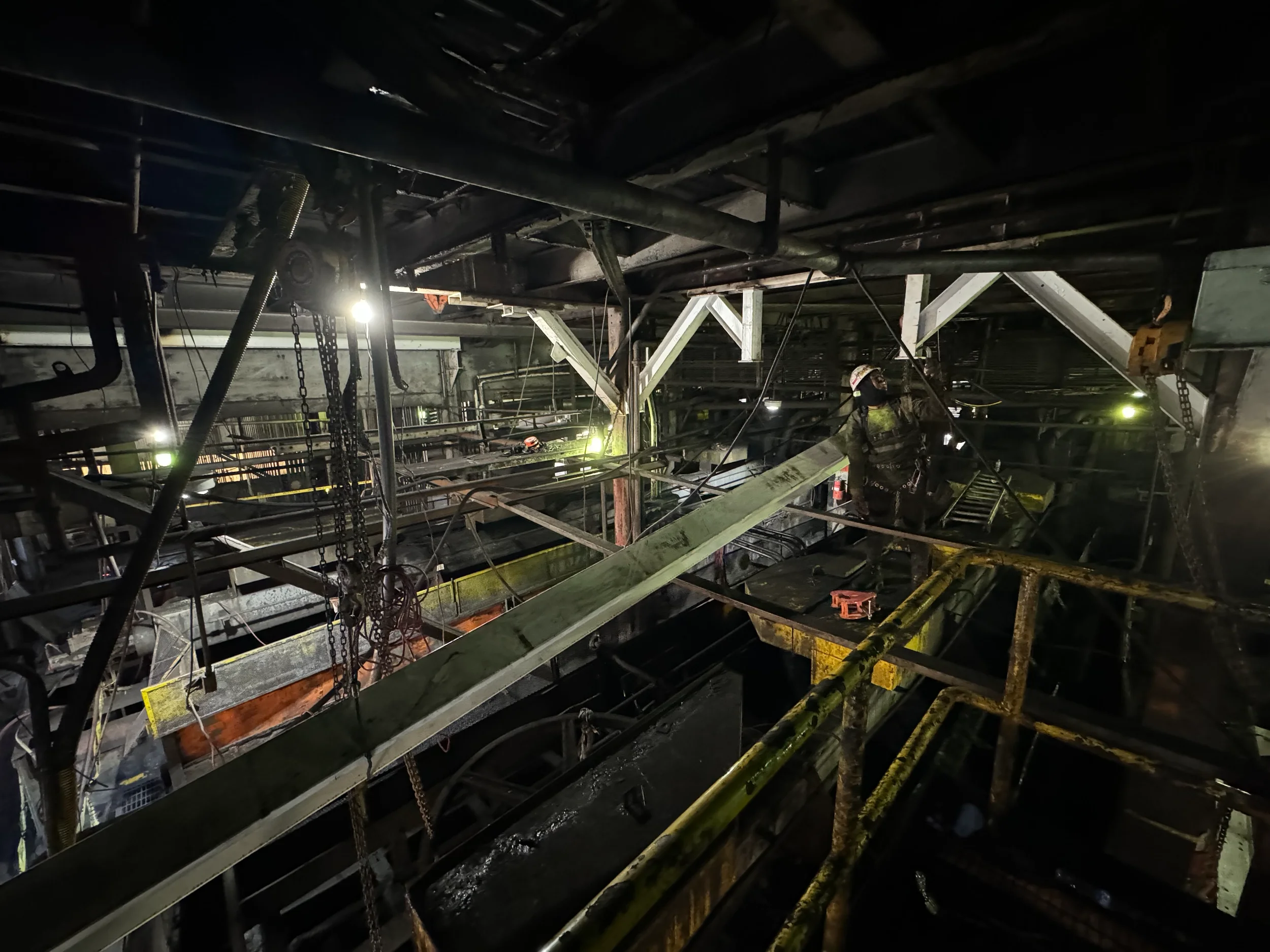 Industrial construction site inside a large building with metal framework, pipes, chains, and a worker wearing a helmet and safety gear.
