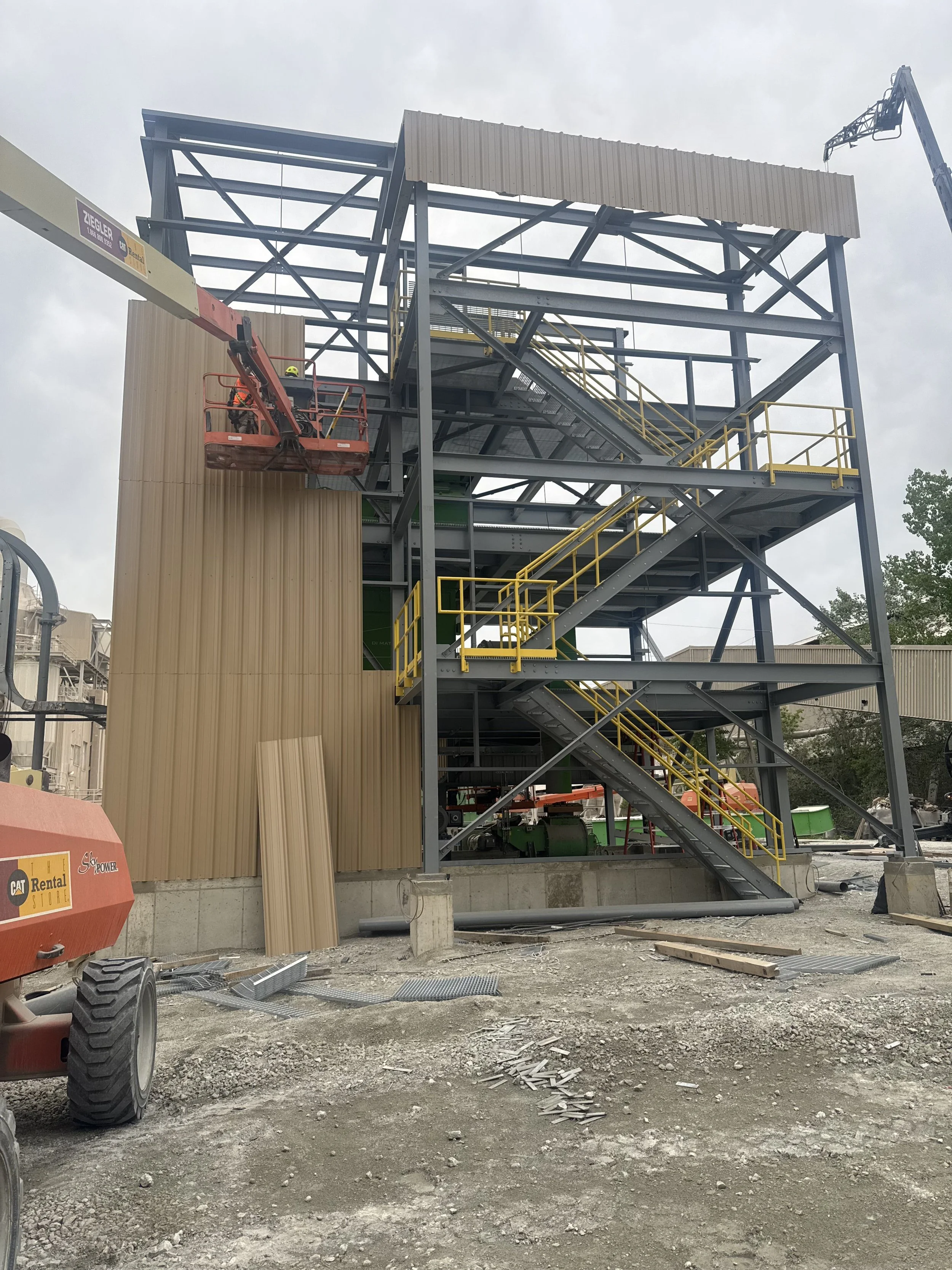 Construction site with a partially built metal structure, stairs, and yellow safety railings. Construction equipment is visible, and the ground is dirt and gravel.