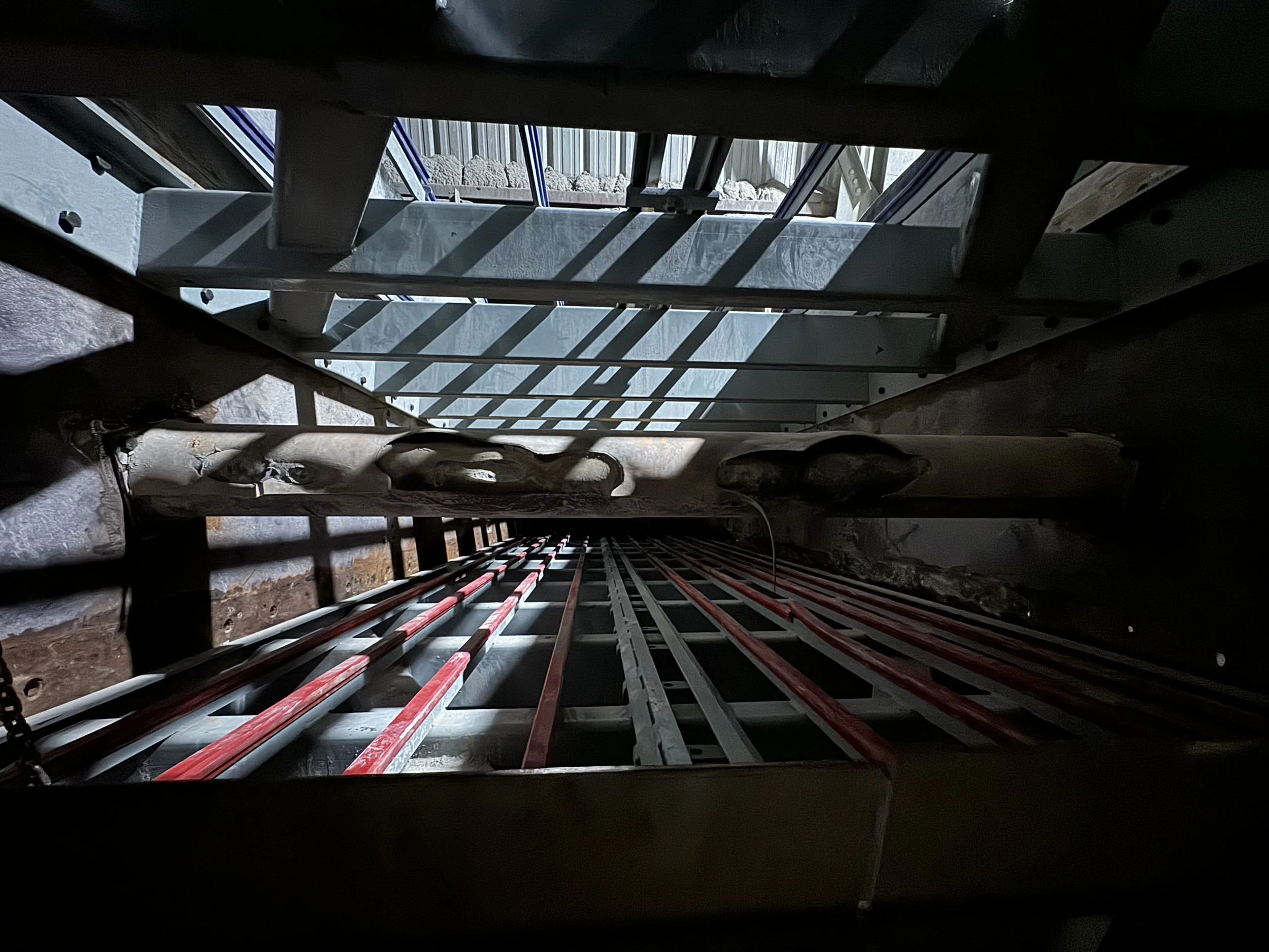 Looking down through a narrow opening in a roof or ceiling, there are metal and wooden structural elements, with sunlight casting shadows from the roof support beams onto the surfaces below.