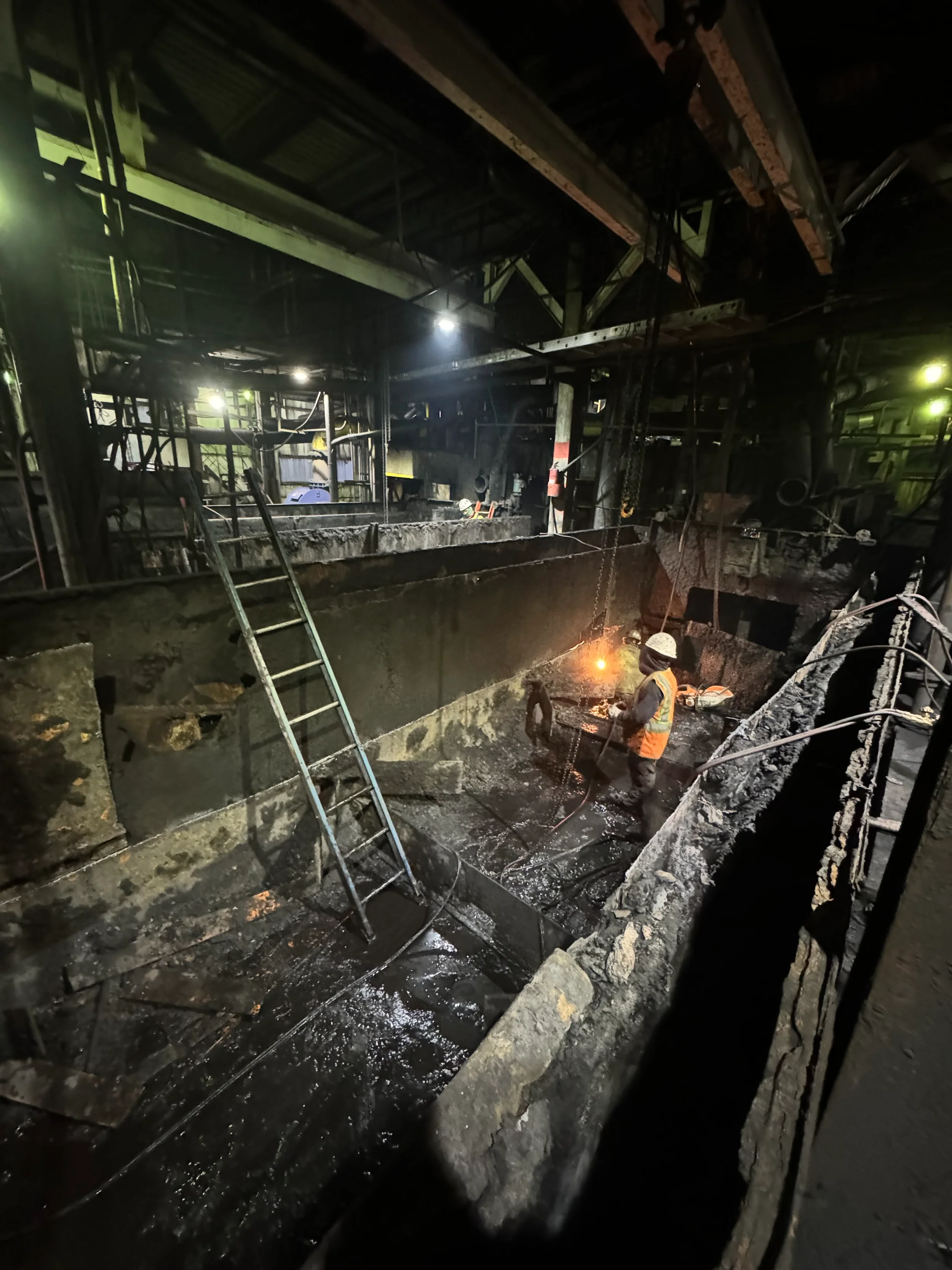 Workers in a coal mine working beneath exposed metal beams and machinery, with dark walls and bright lights illuminating the underground space.