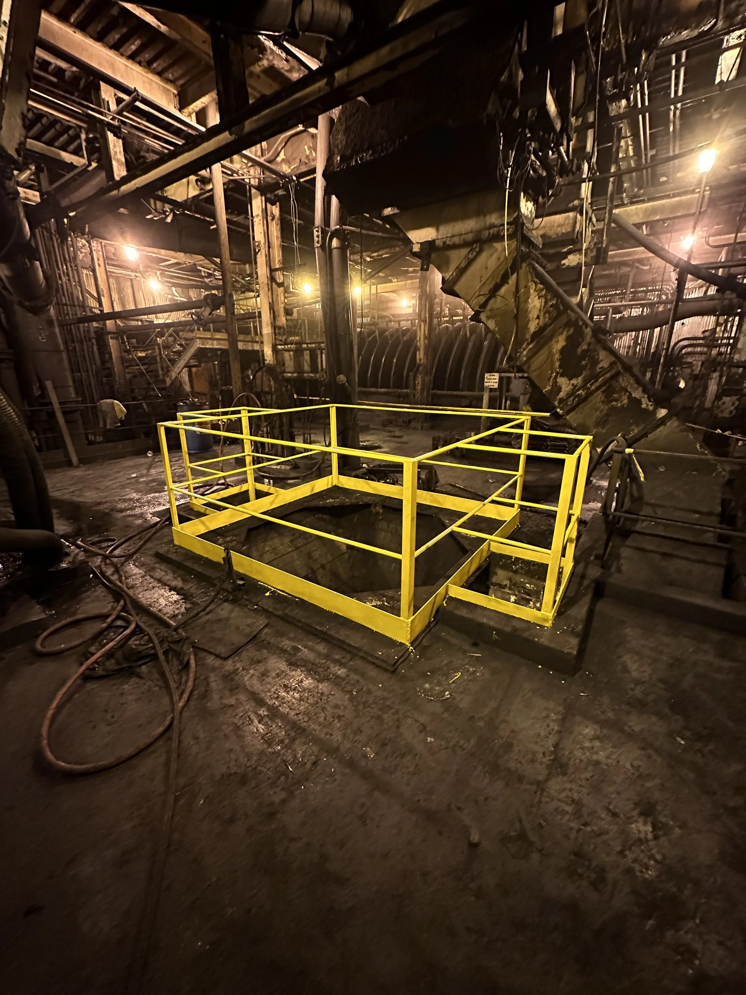 Interior of a factory or industrial facility with exposed pipes, machinery, and a yellow safety railing around a hazardous area. The space appears greasy and dark, with warm lighting.