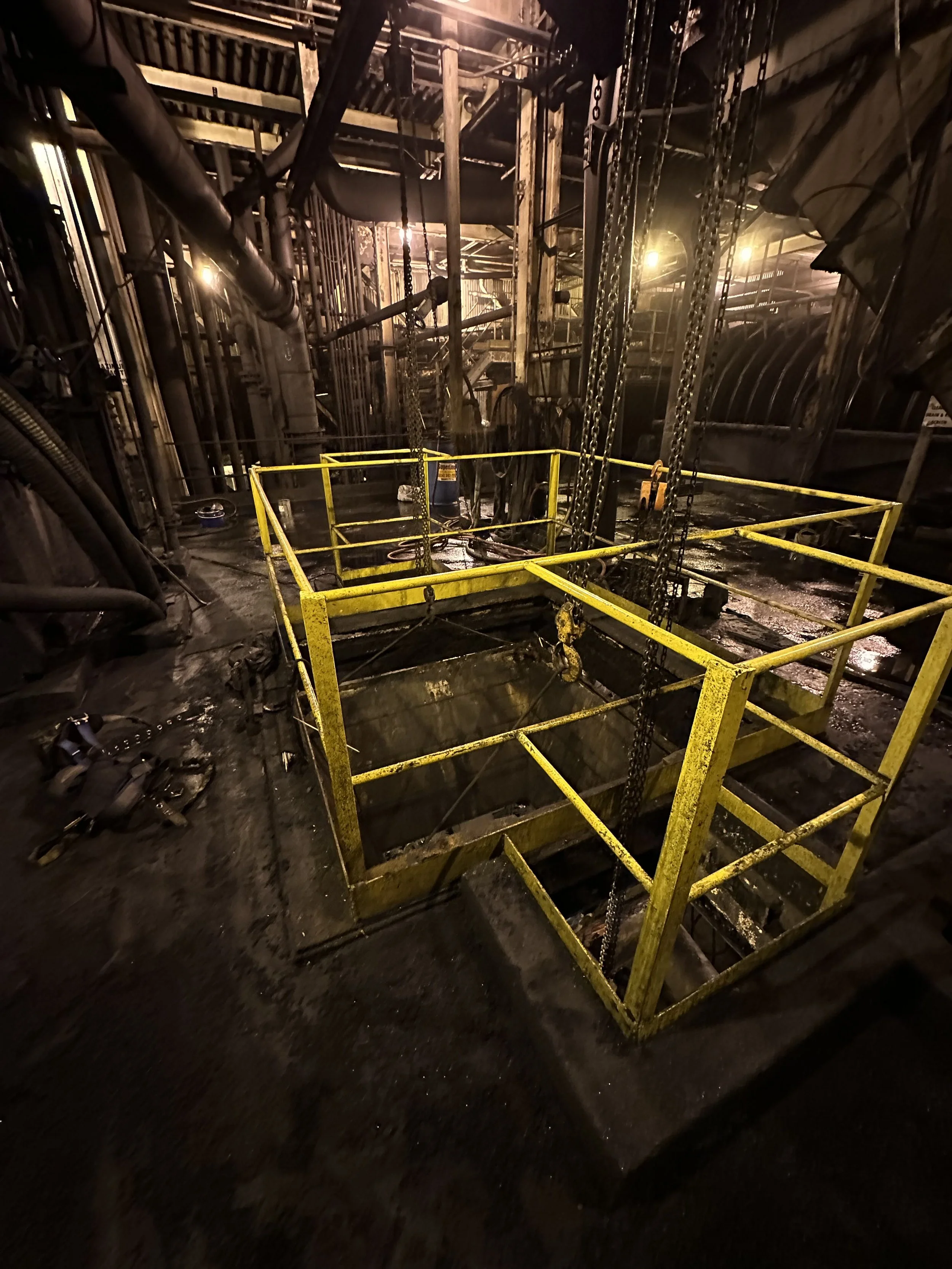 Interior of an industrial or utility space with metal pipes, chains, and yellow safety railing around a pit, with dim lighting.