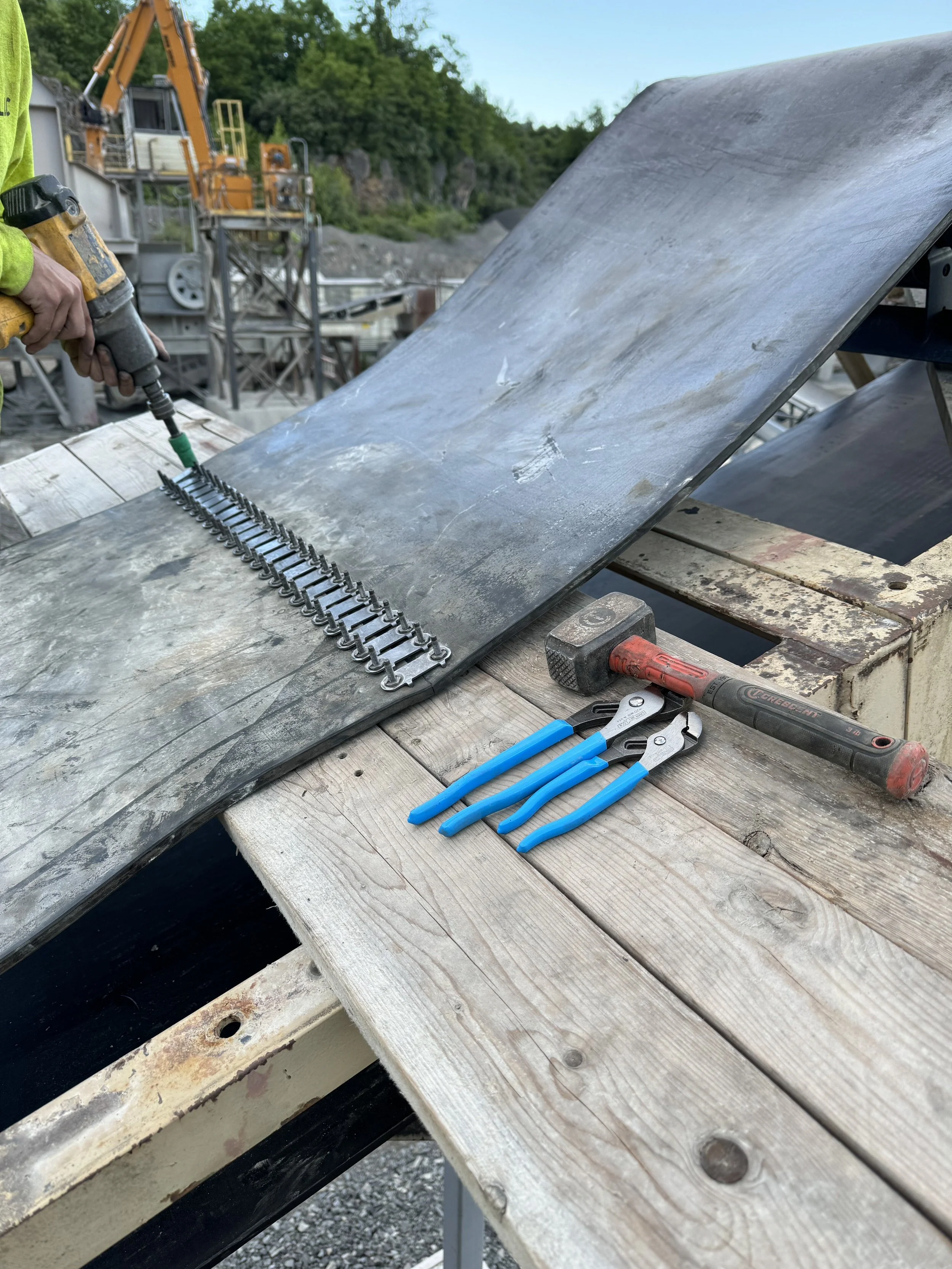 A person working on a metal construction project outdoors, using a power drill, with tools such as pliers and a hammer on a wooden work surface, and a large curved metal sheet.