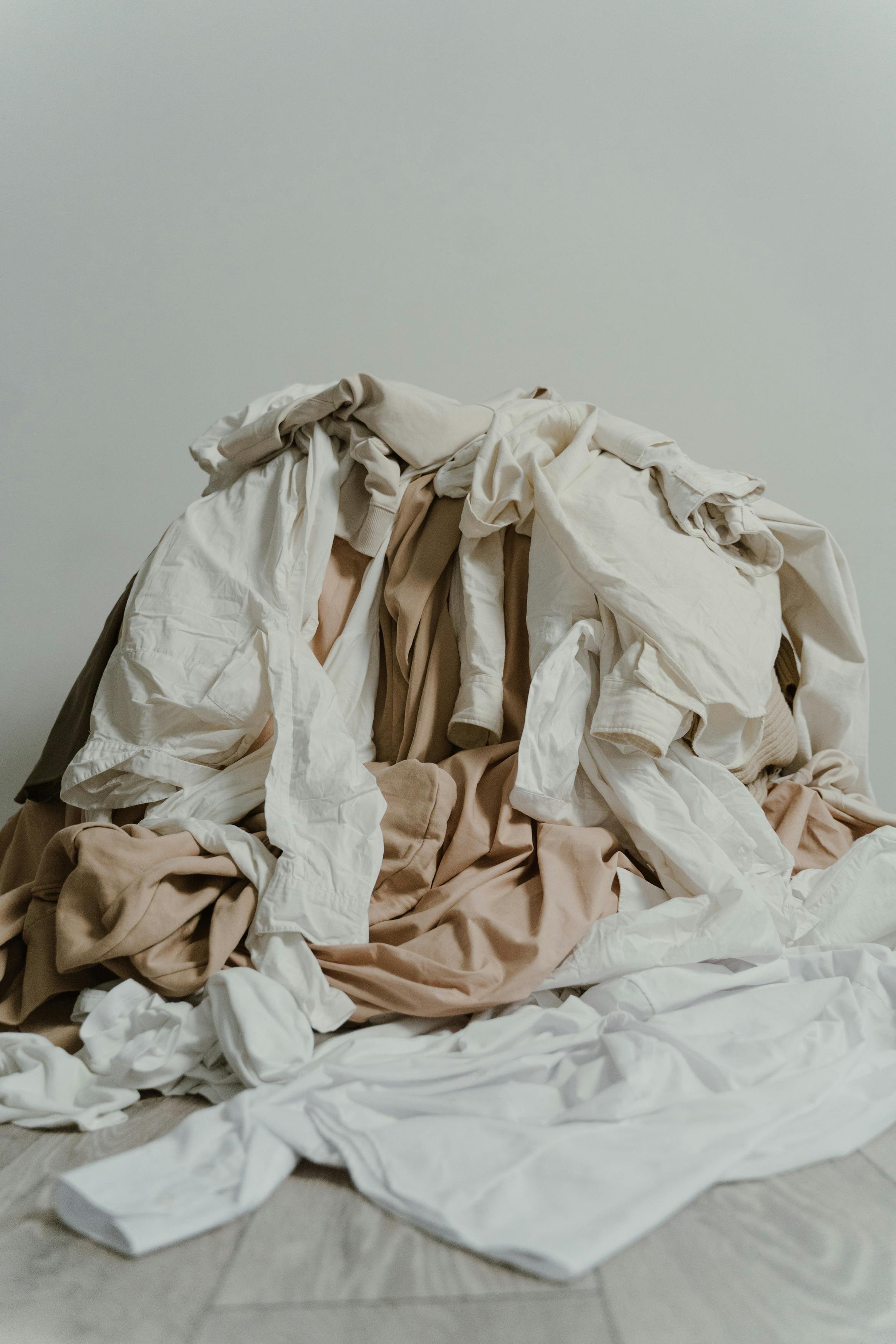 A pile of tangled, wrinkled, and crumpled white, beige, and brown cloth or fabric on a light-colored floor in front of a plain white wall.