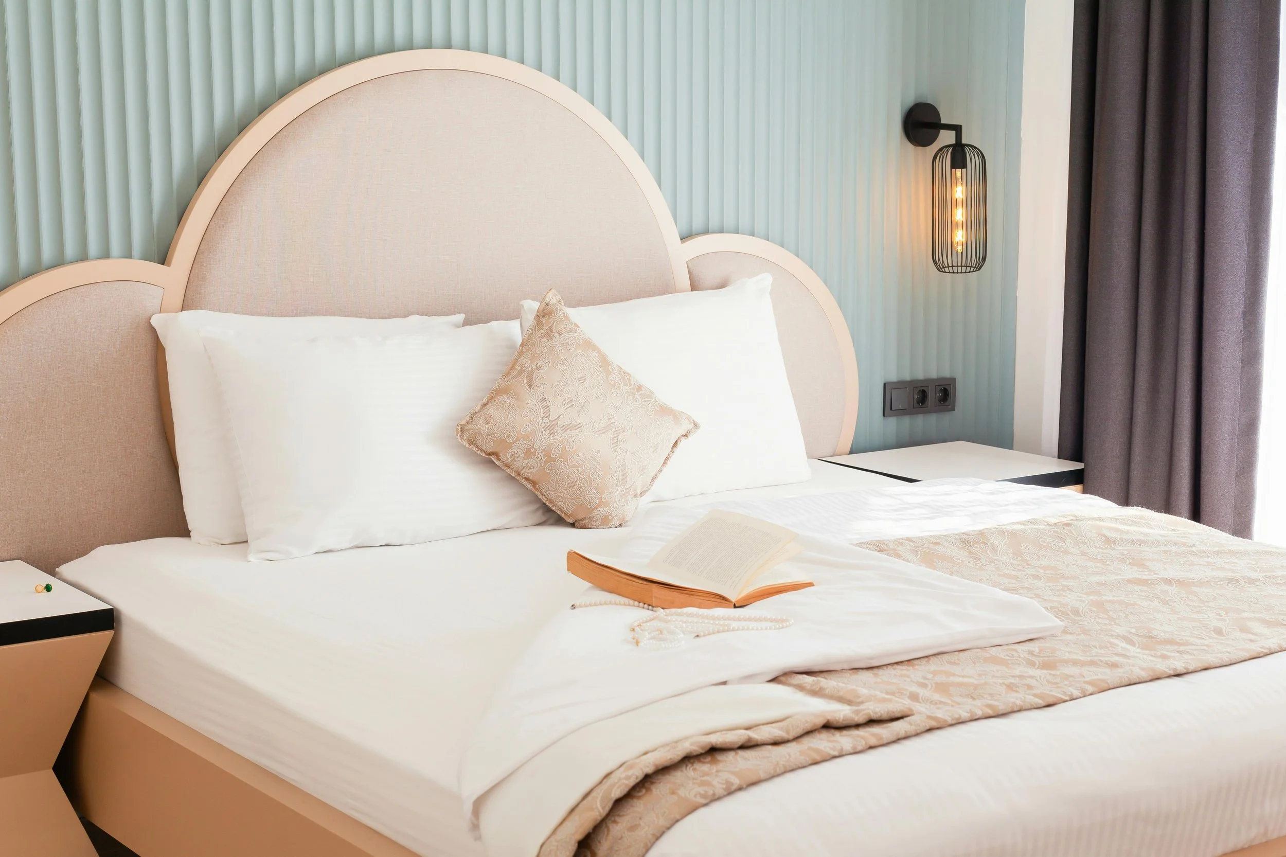 A neatly made bed with white linens, a beige decorative pillow, and an open book resting on the bedspread. The bed has a modern headboard, and behind it is a textured, light blue wall with vertical pleats. To the right, there is a black wall-mounted light fixture with an exposed bulb and dark curtains partially covering a window.