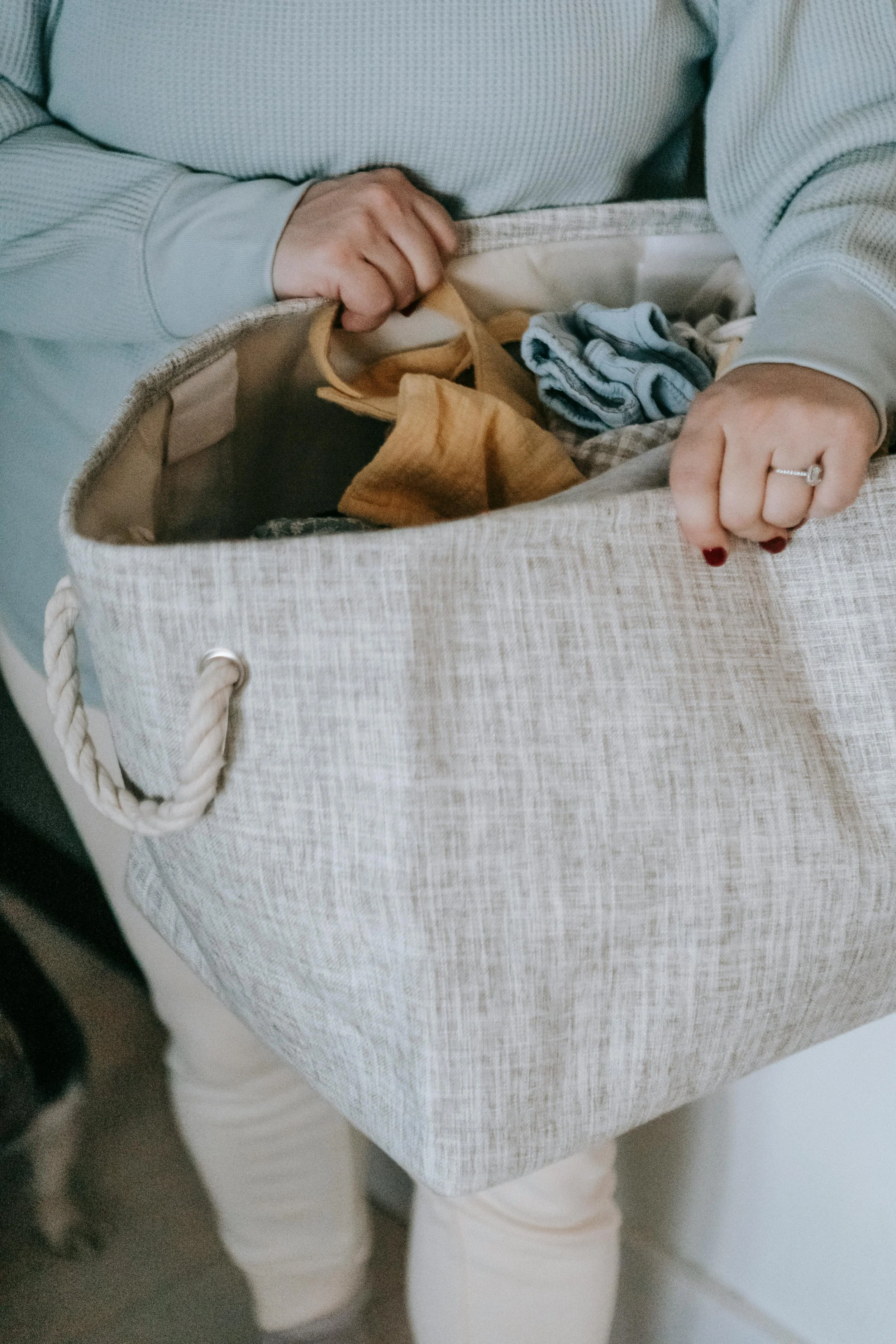 Person in a light-colored, long-sleeve shirt and white pants is reaching into a woven beige tote bag with rope handles, which contains clothing items, including a pair of striped socks.