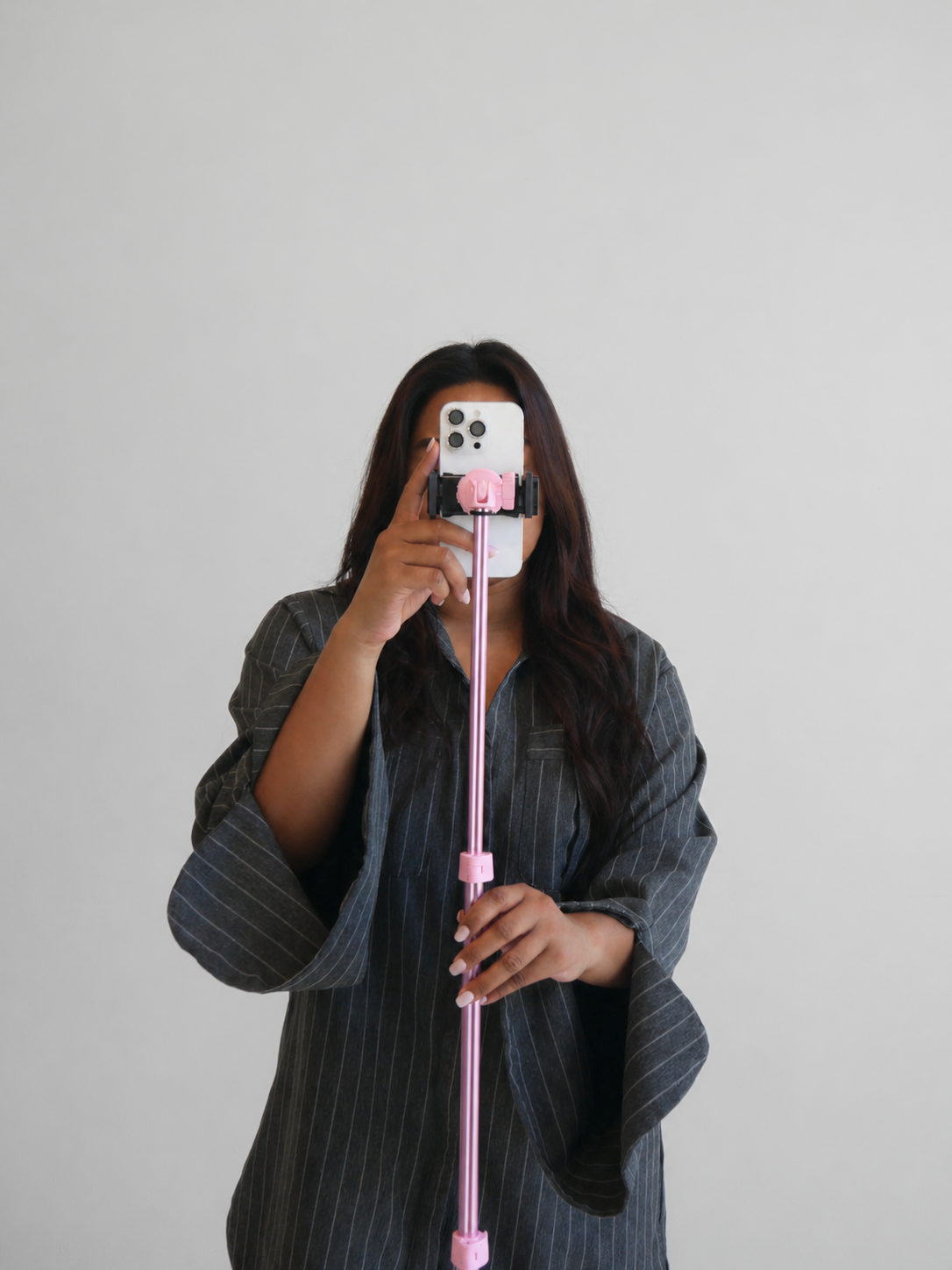 A woman with long dark hair wearing a gray pinstripe shirt takes a selfie using a smartphone mounted on a pink selfie stick against a plain white background.