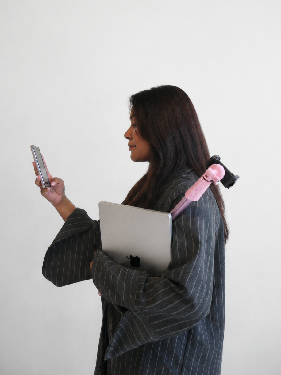 A woman with long dark hair, wearing a pinstriped blazer, holding a silver MacBook in her left arm, and looking at her smartphone with her right hand. She has a pink selfie stick attached to her shoulder.